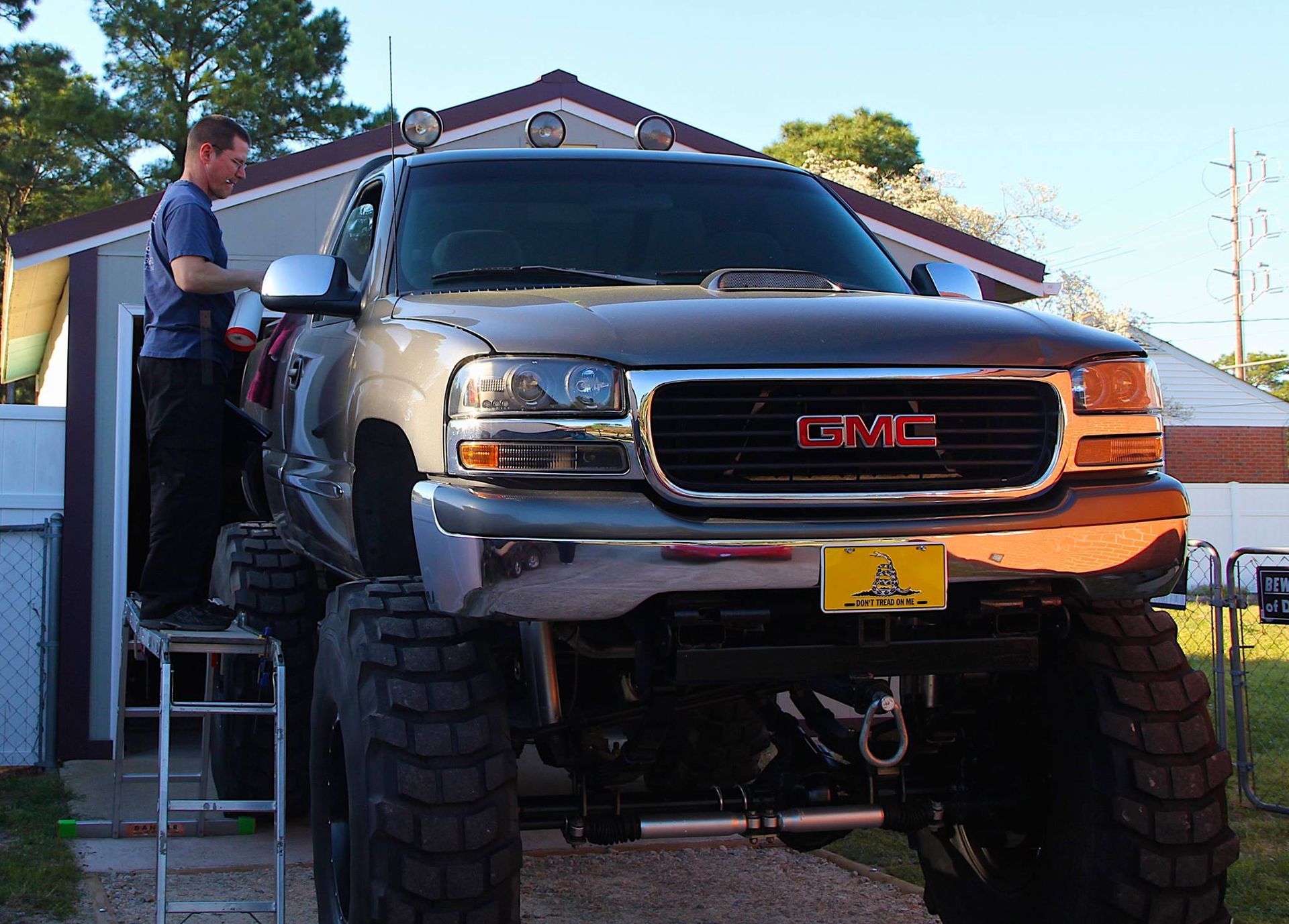 A person stands on a ladder to reach inside the cab of a large lifted gray GMC pickup truck parked in front of a garage.