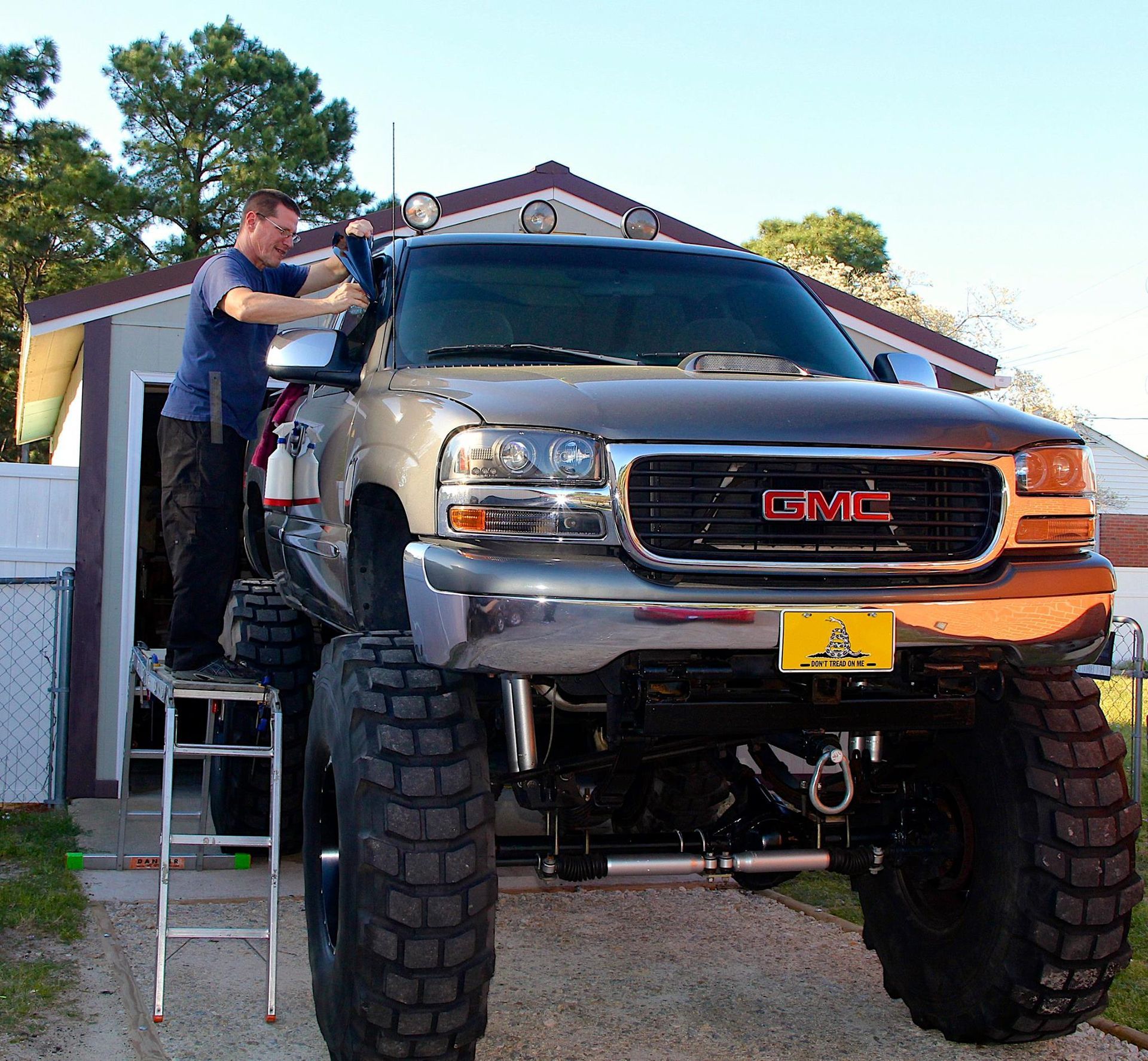 A person stands on a tall ladder to reach the side mirror of a large, lifted silver GMC truck parked outdoors.