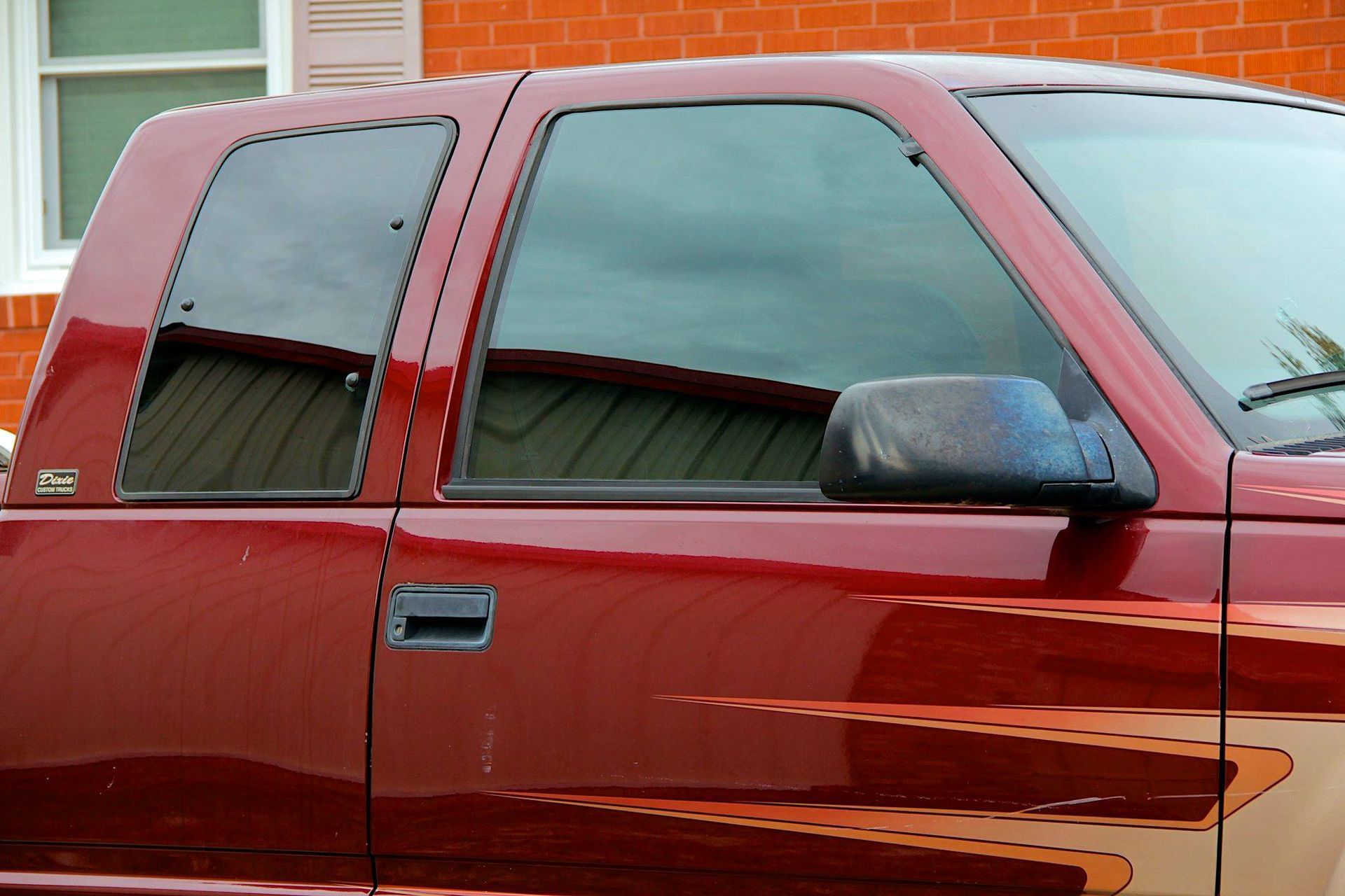 A close-up side view of a maroon pickup truck with tinted windows, parked in front of a brick building.