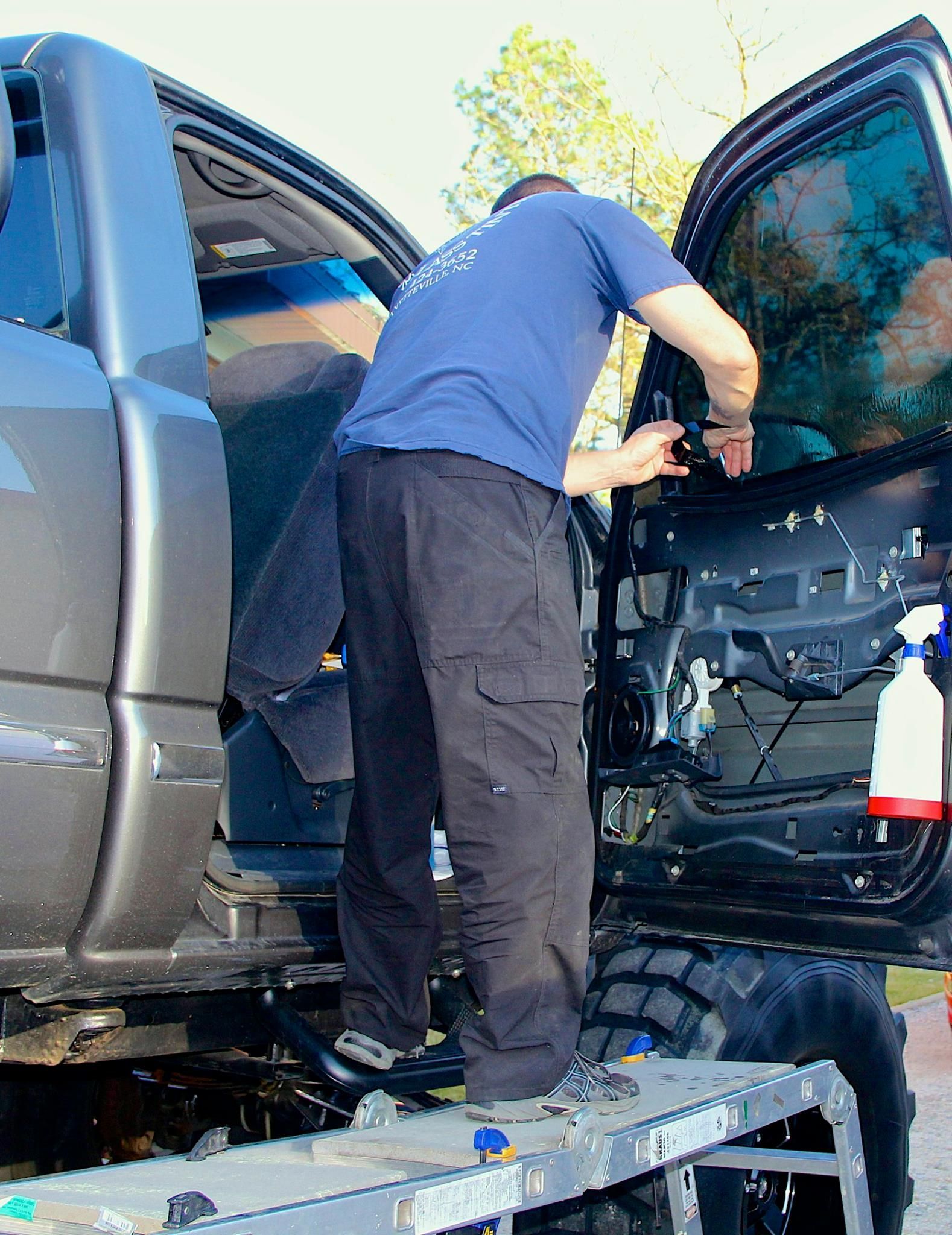 A person stands on a step ladder, working inside the open door panel of a dark grey pickup truck.