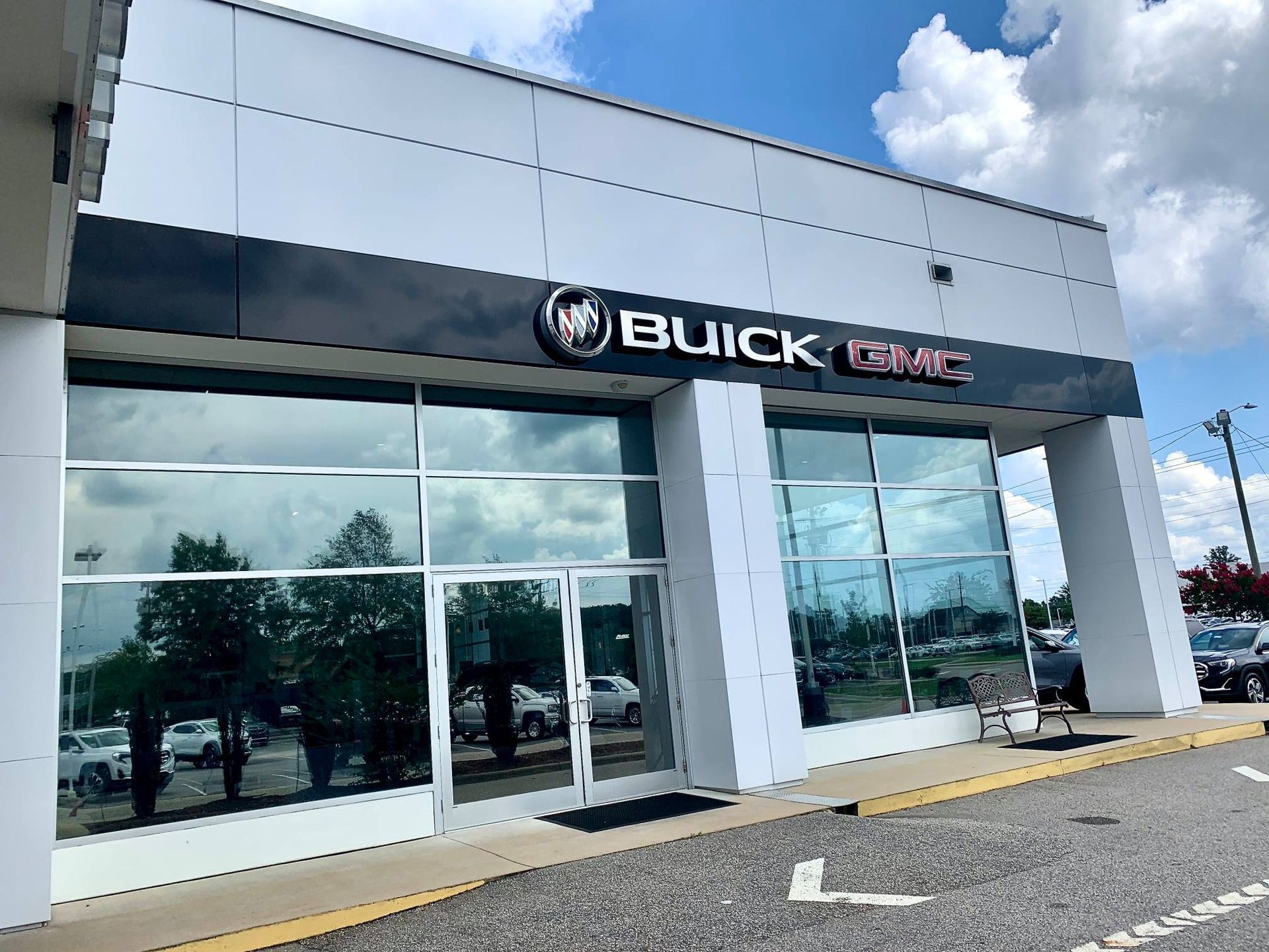 A modern white Buick GMC car dealership building with large glass windows under a blue, cloudy sky.