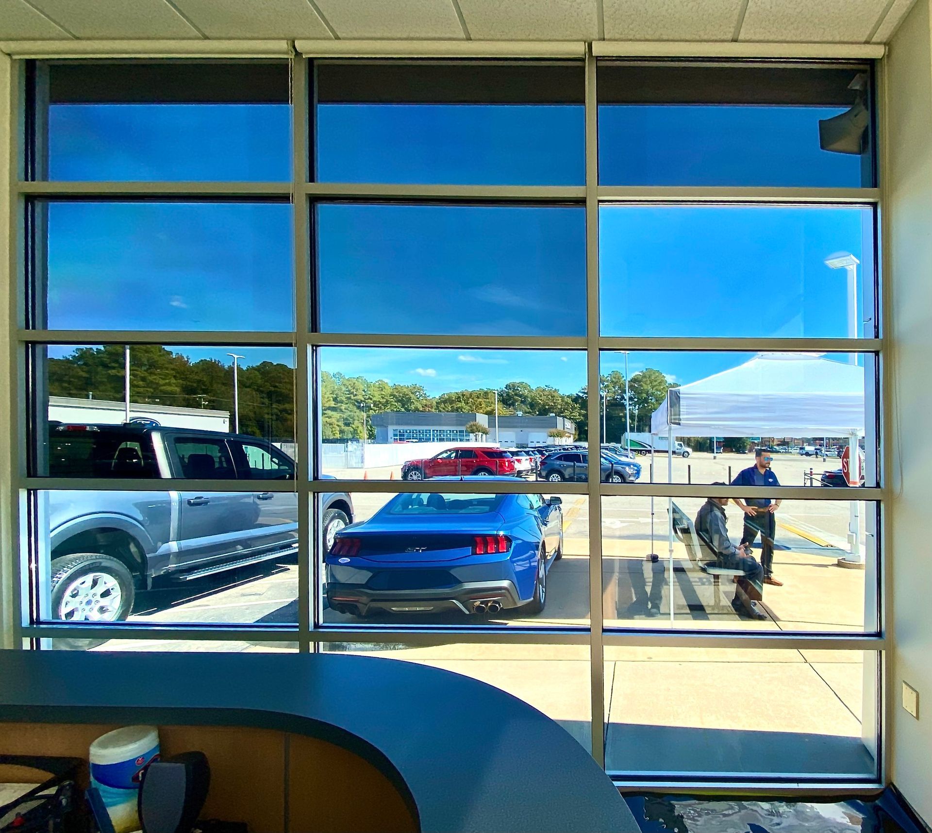 A blue Ford Mustang and a grey pickup truck are parked outside a dealership, visible through a large, paneled window.