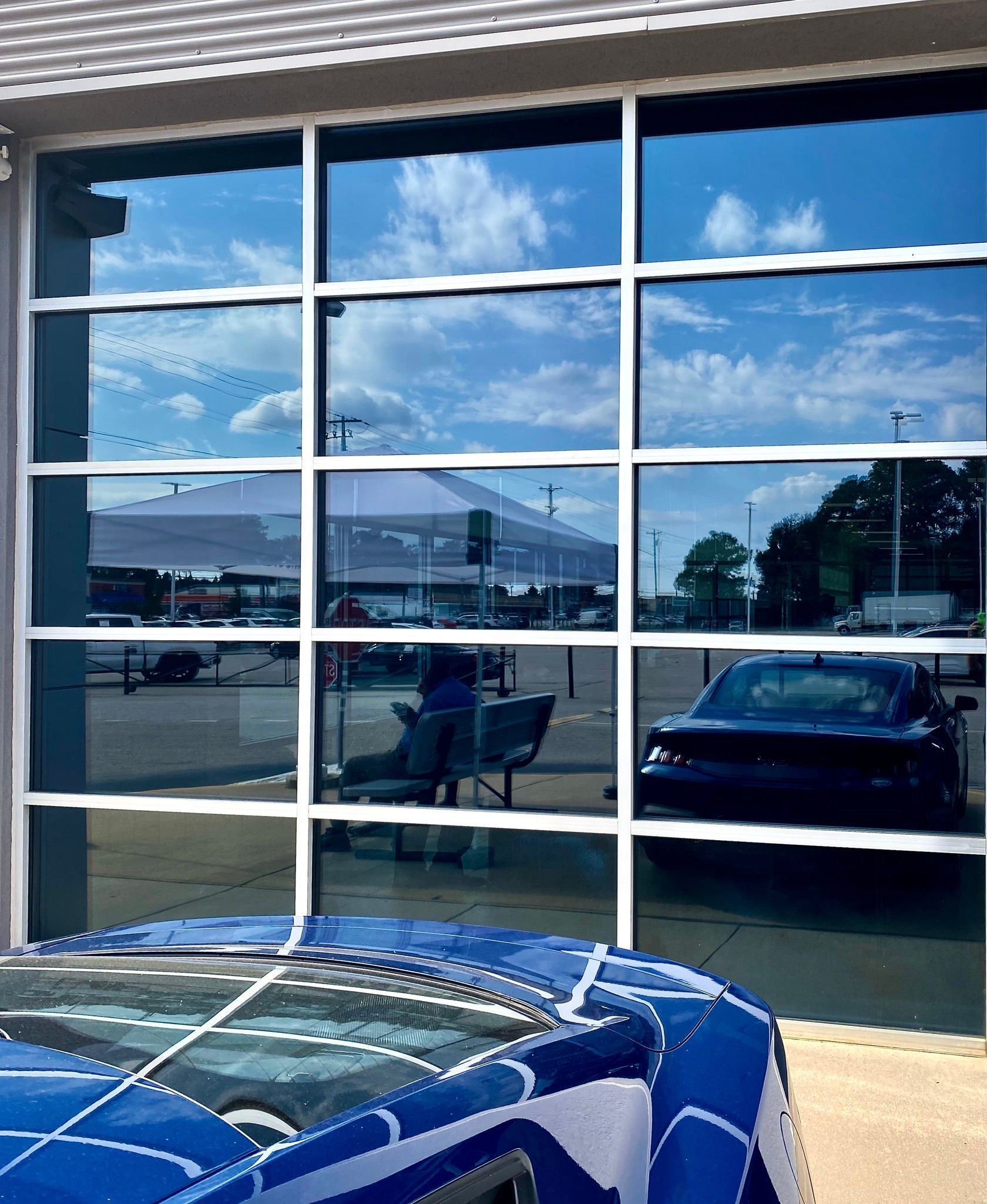 A blue car cover with a white grid pattern sits outside a building with glass panels reflecting a parking lot and a car.