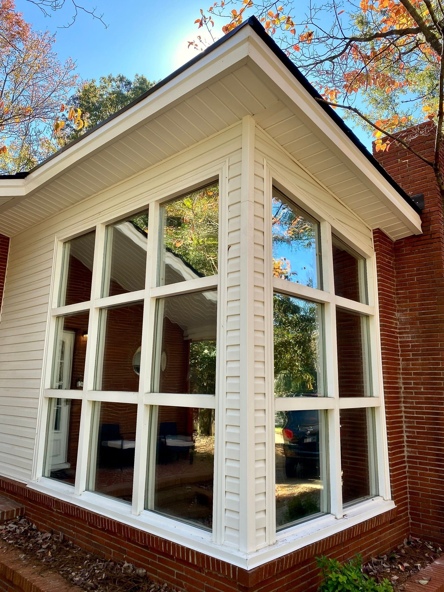 A sunroom corner with white-framed, grid-style glass walls set against red brick, bathed in autumn sunlight.
