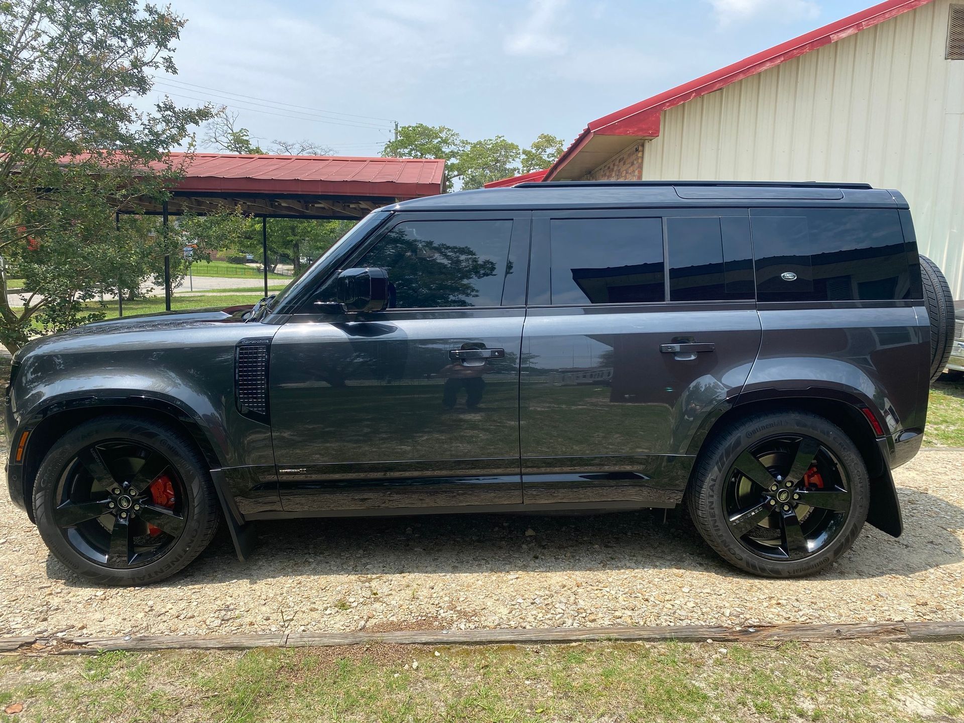 A side view of a dark grey, modern Land Rover Defender SUV parked on a gravel lot near buildings.