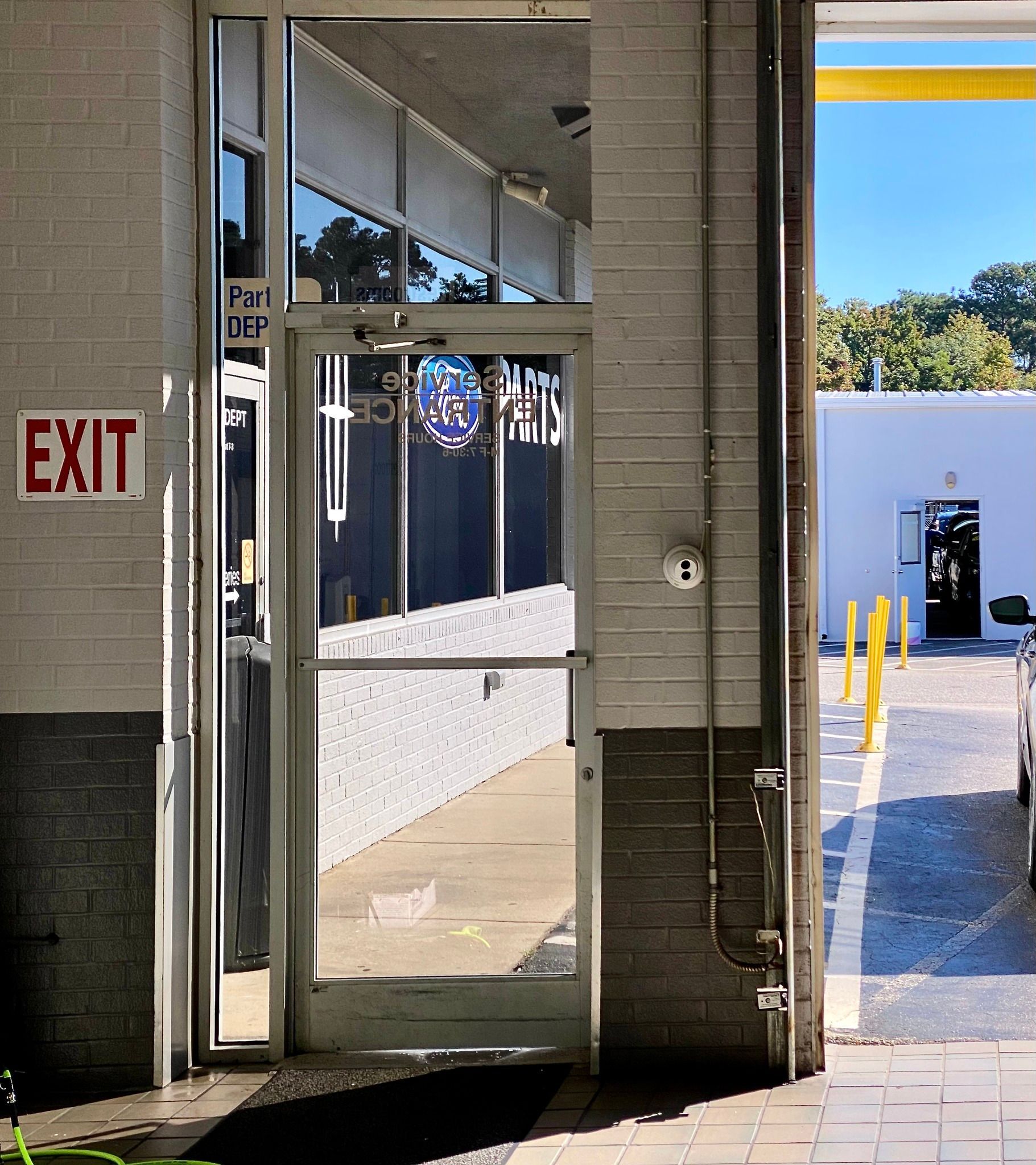 A glass door set in a white brick wall next to an exit sign, looking out to an outdoor path and another building.