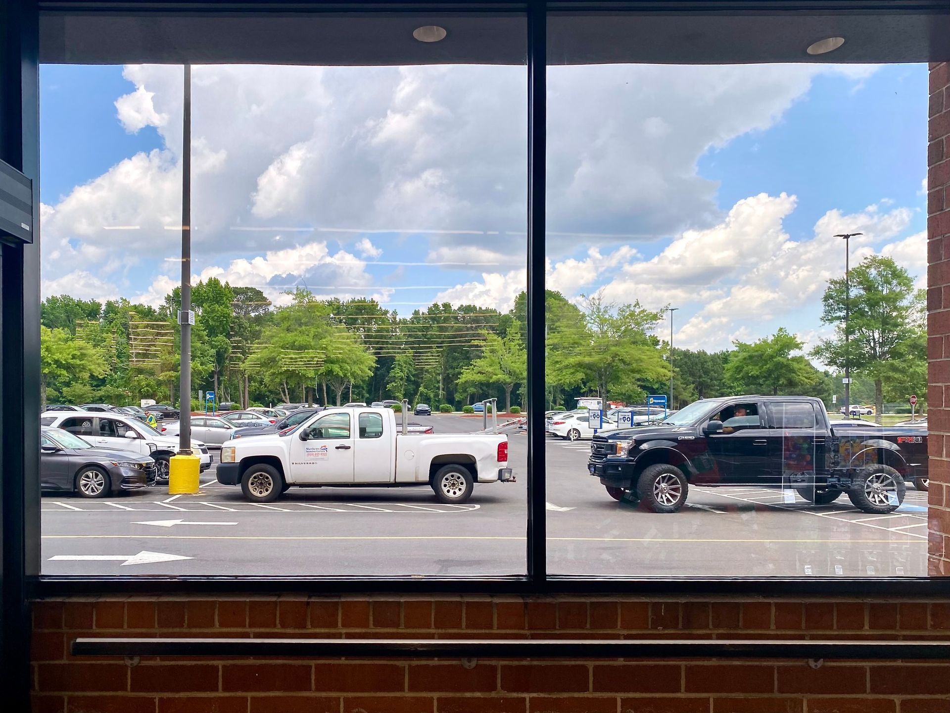A parking lot view through a window, showing a white pickup truck, a black lifted pickup truck, and several parked cars.