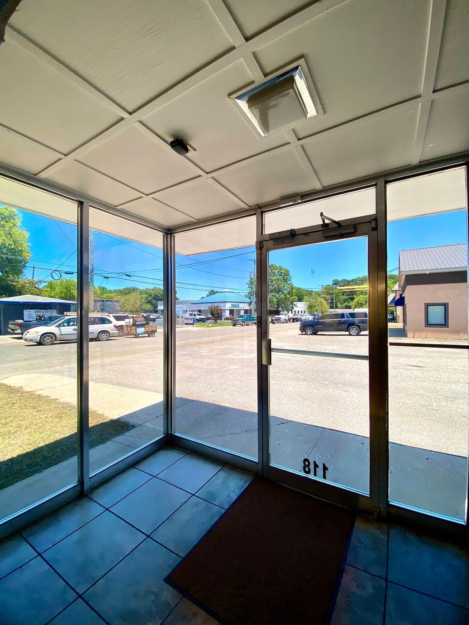 A view from inside a building looking out through a glass-walled entrance onto a parking lot on a sunny day.