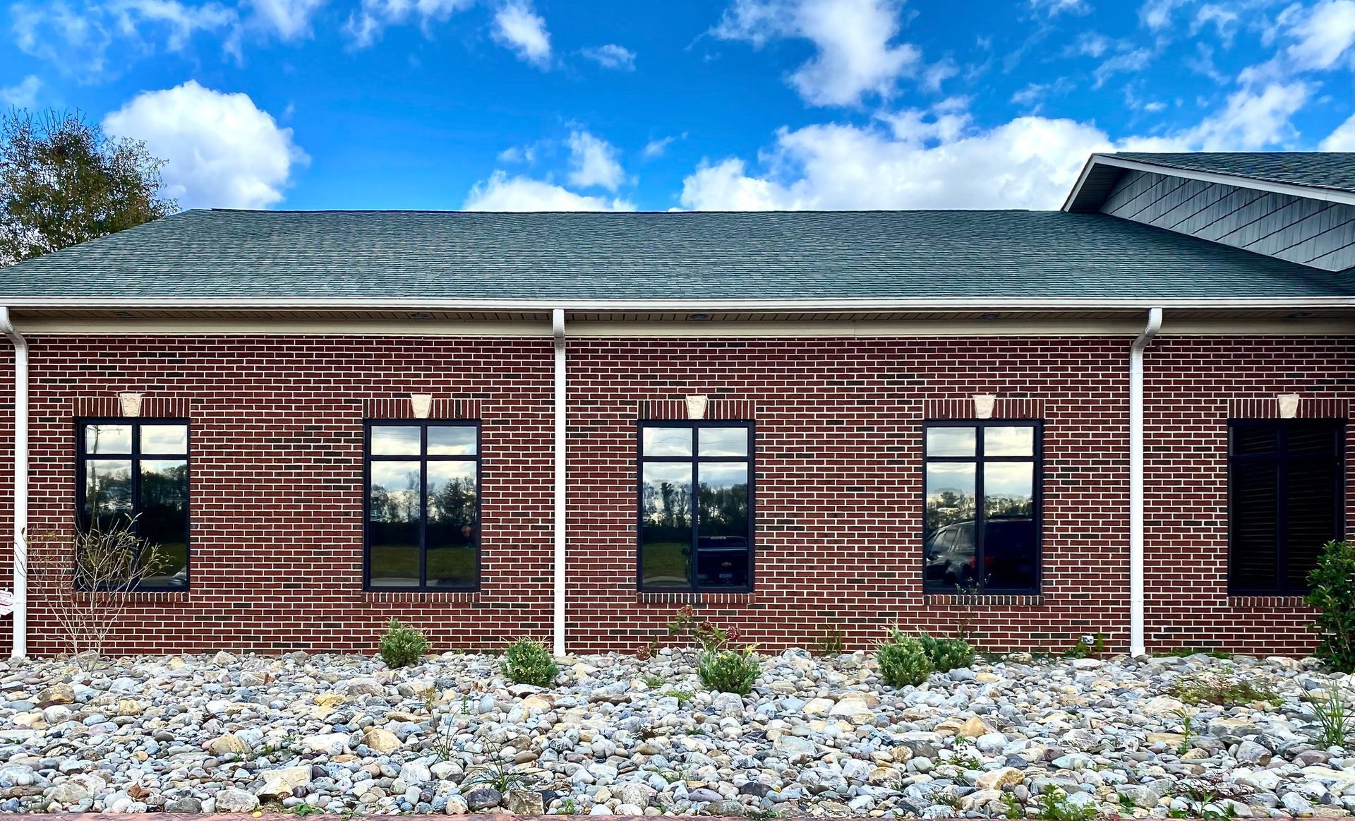A single-story brick building with five black-framed windows, a gray shingled roof, and a gravel base under a blue sky.