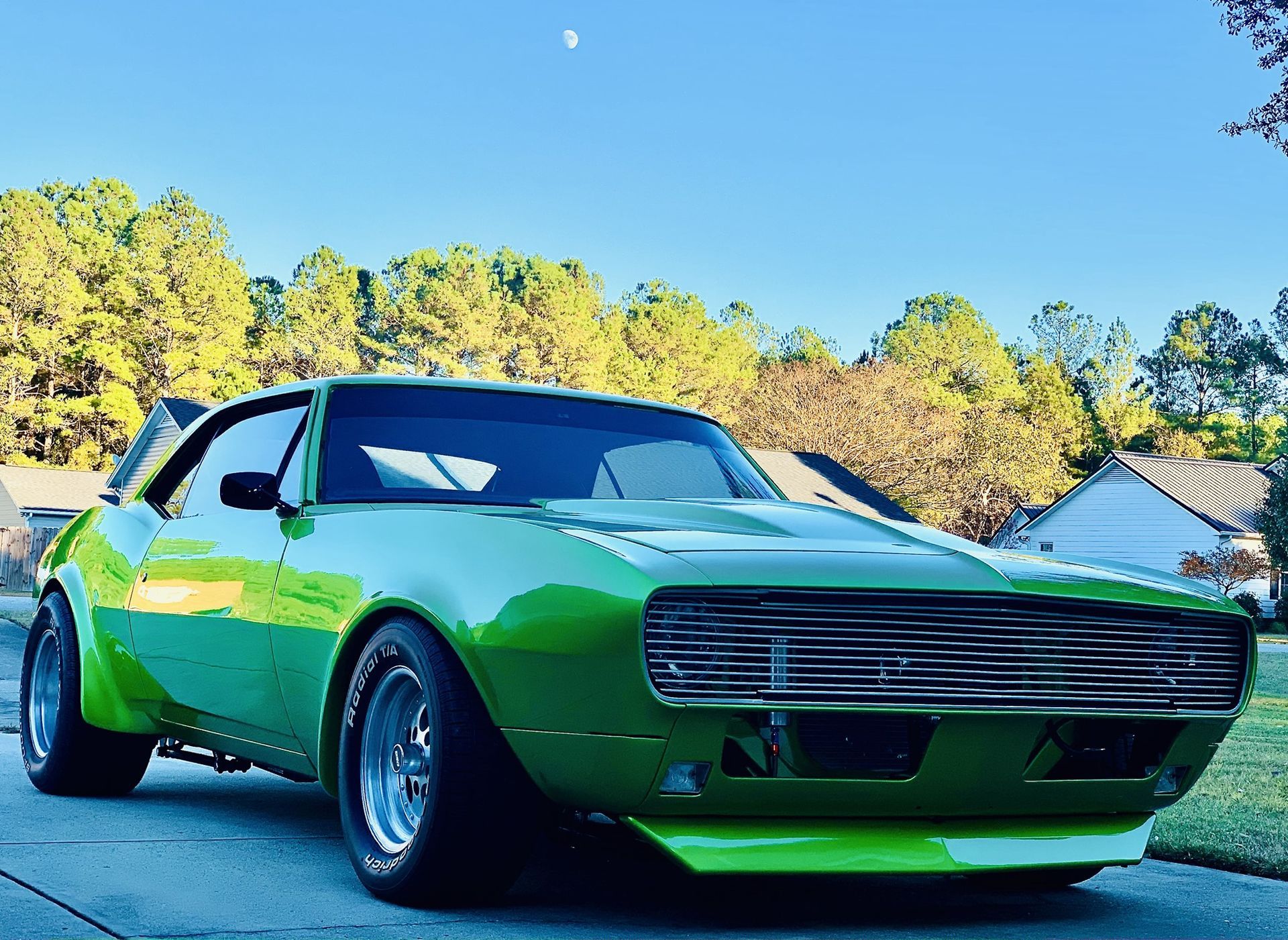 A bright green, modified vintage Chevrolet Camaro parked on a driveway with trees and houses in the background.
