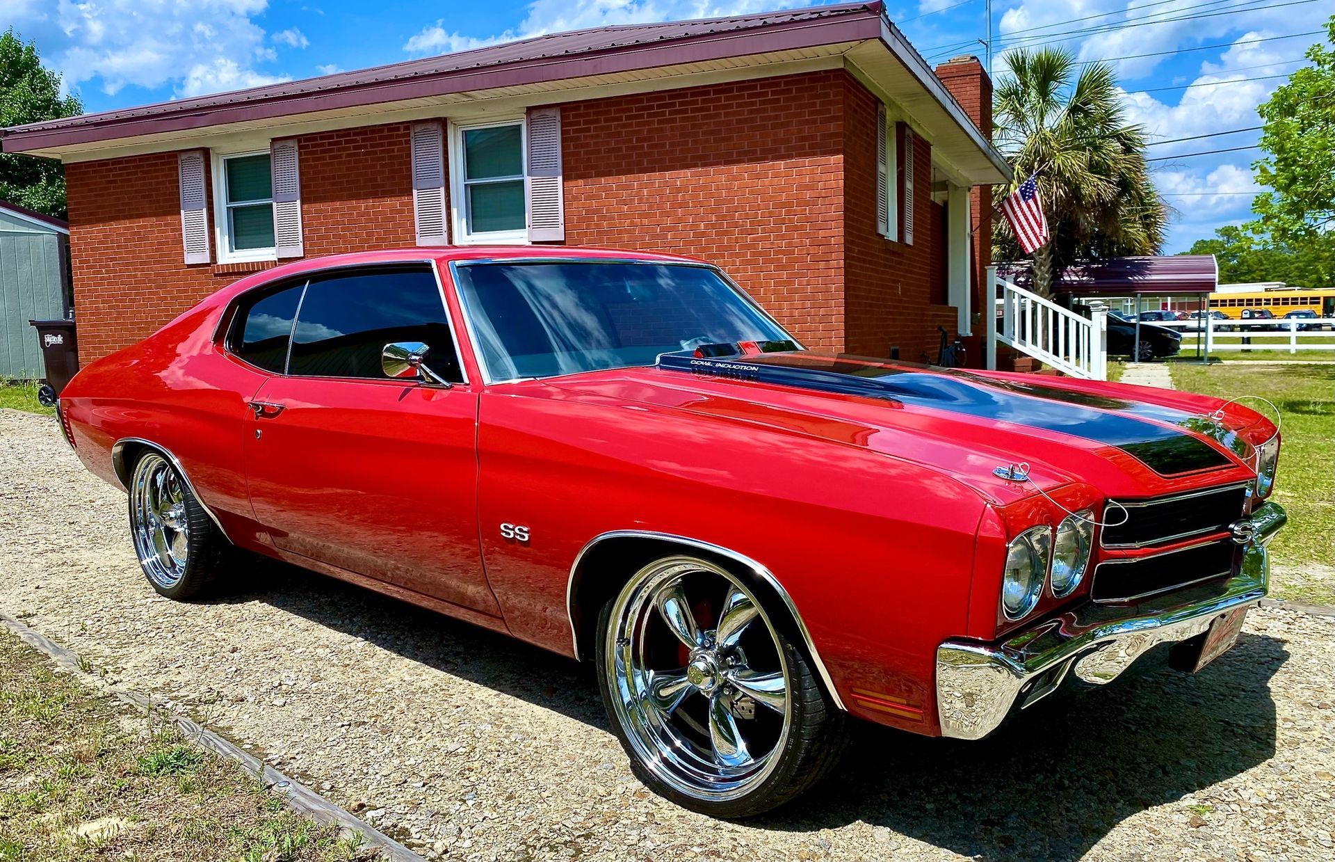 A shiny red 1970 Chevrolet Chevelle SS parked on a gravel driveway in front of a red brick house under a blue sky.