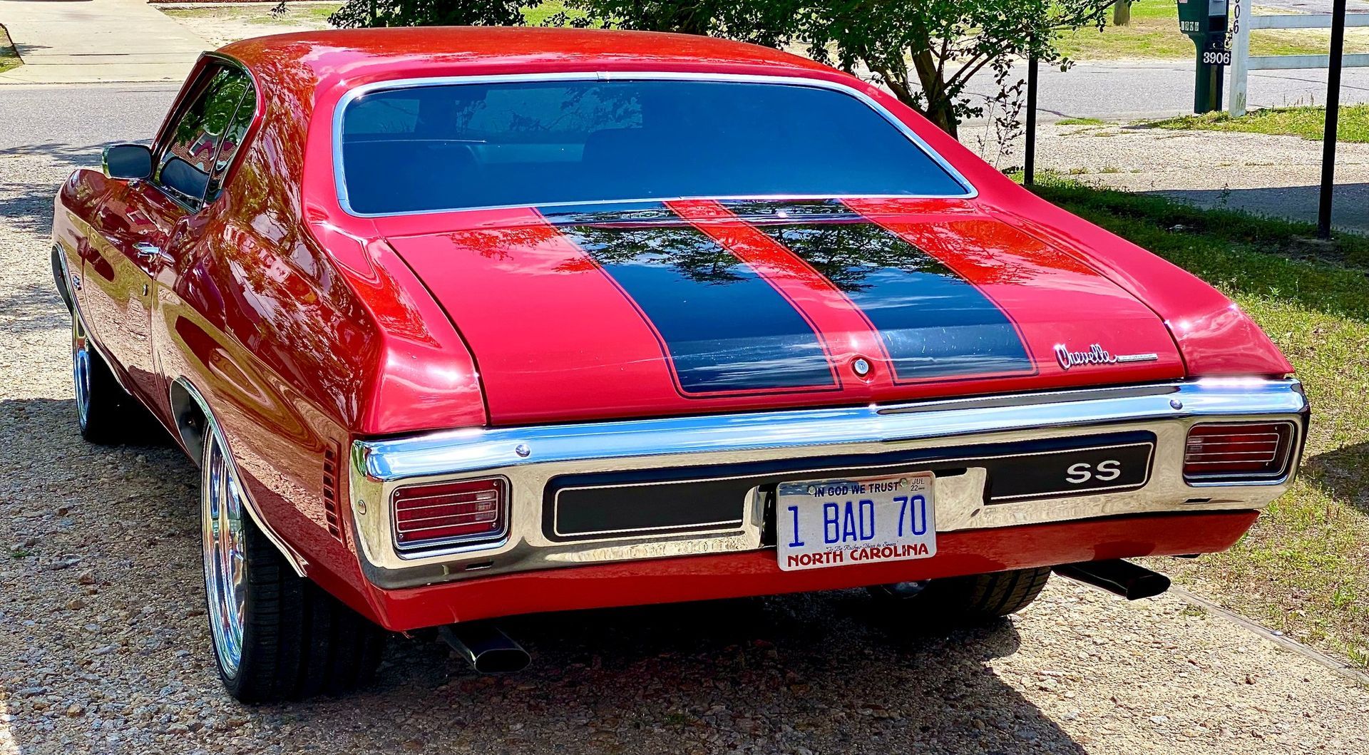 A red vintage Chevrolet Chevelle with black racing stripes parked on a gravel surface.