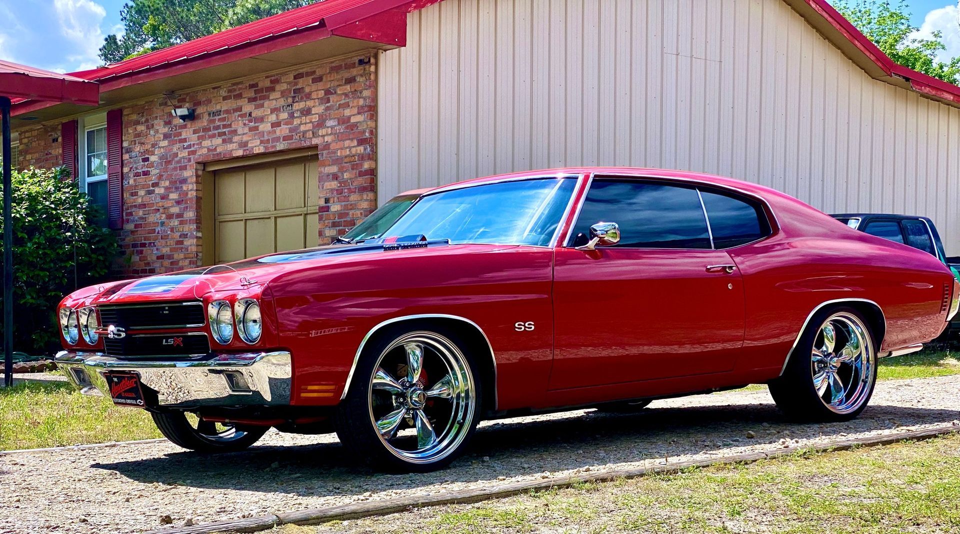 A red classic Chevrolet Chevelle parked on a gravel driveway in front of a building.