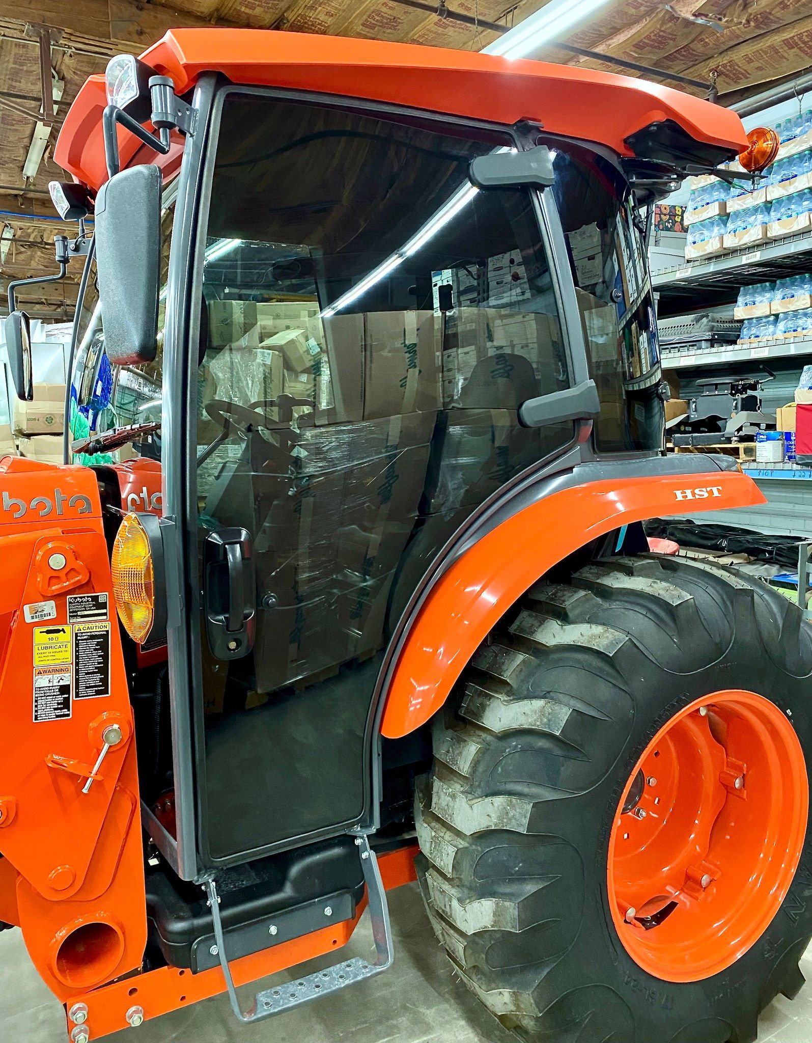 An orange Kubota tractor cabin with a door, side mirror, and large tire, parked inside a well-stocked workshop.