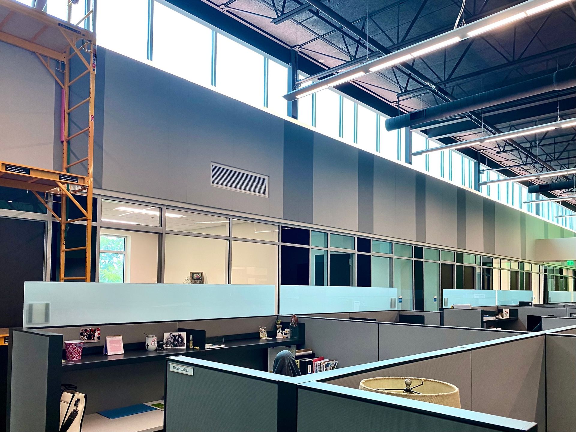 A modern office space features rows of cubicles under high windows and industrial ceilings with a scaffolding unit nearby.