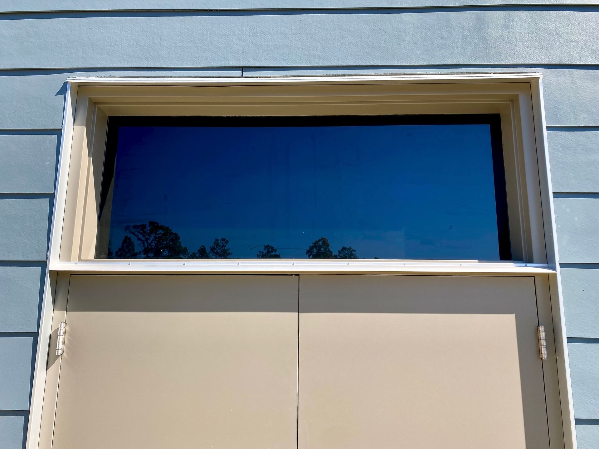 A rectangular window with dark glass, framed in beige, set into a light blue horizontal-sided wall.