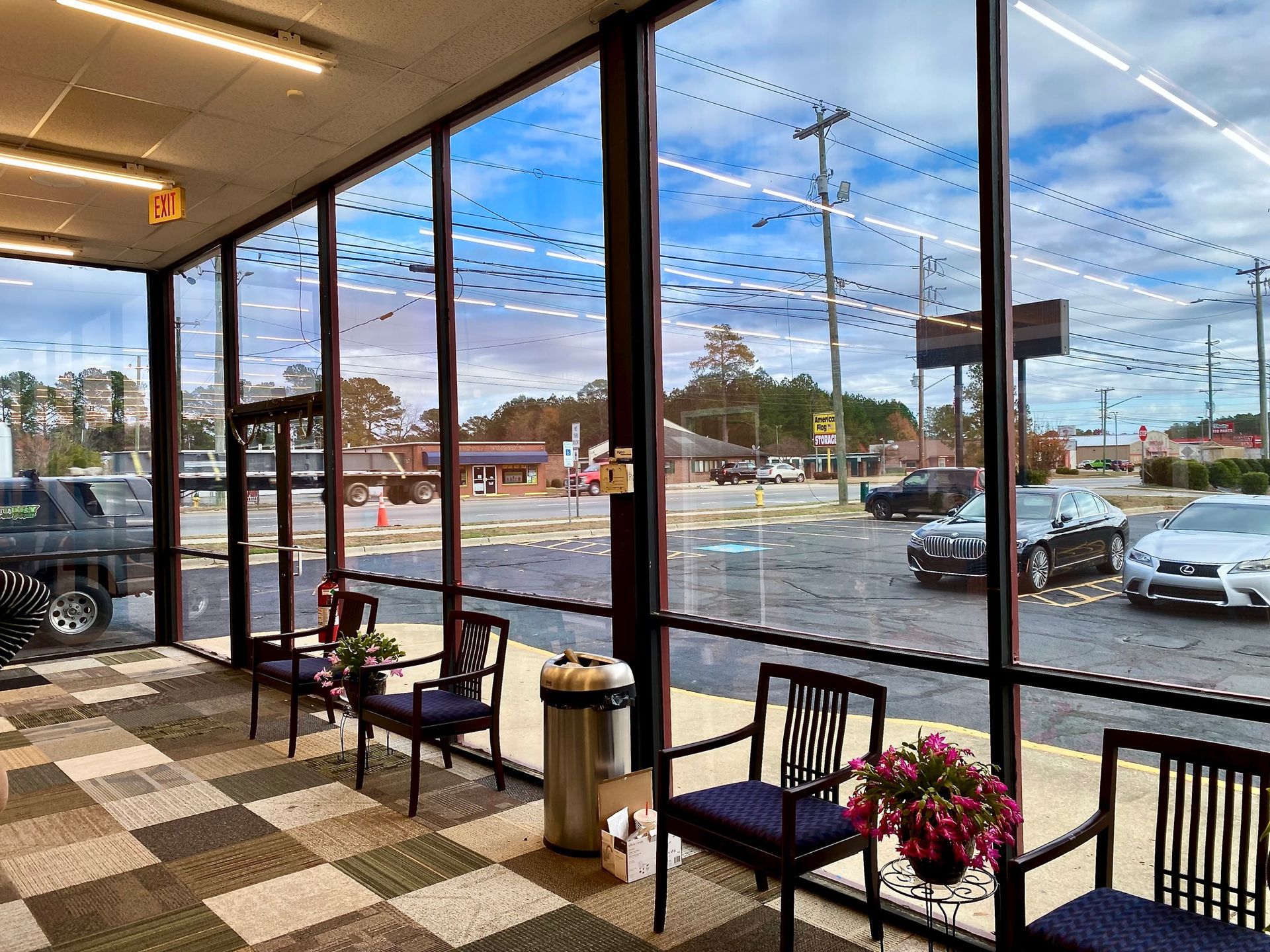 A bright indoor seating area with blue chairs and a trash can, overlooking a parking lot through large glass windows.
