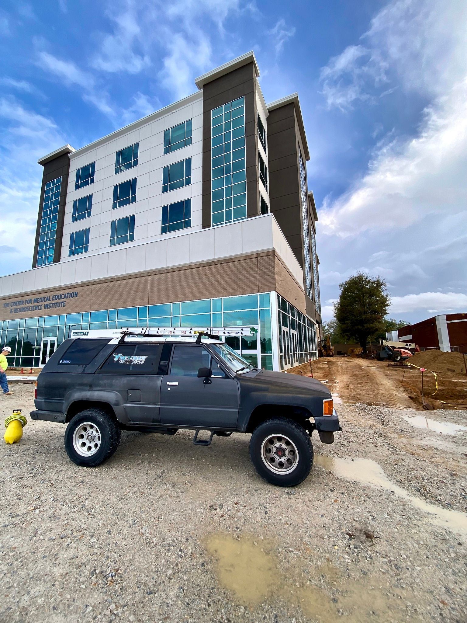 A dark gray Toyota 4Runner parked on a gravel lot in front of a modern, multi-story building under construction.