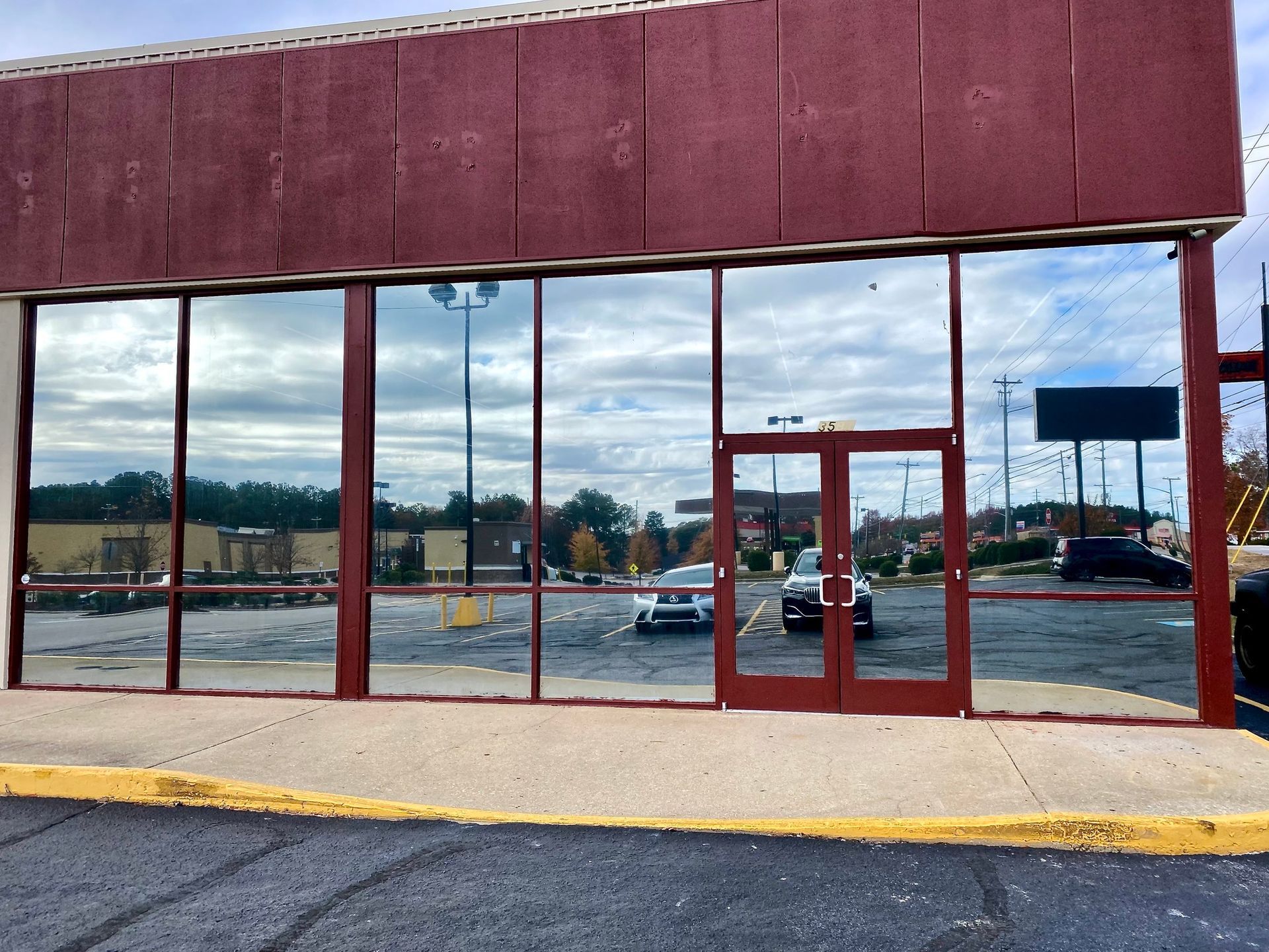 A storefront with large glass windows and a red metal frame, reflecting a parking lot and a cloudy sky.