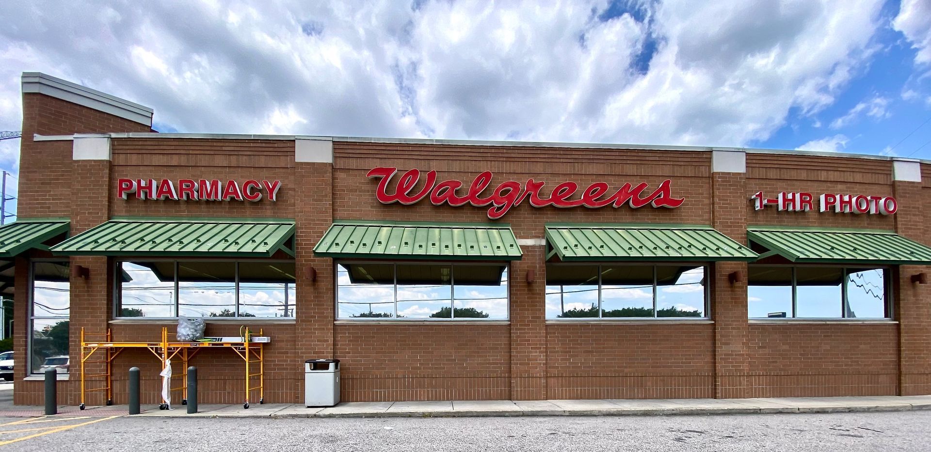 The brick storefront of a Walgreens pharmacy featuring red signage, green awnings, and large, reflective glass windows.