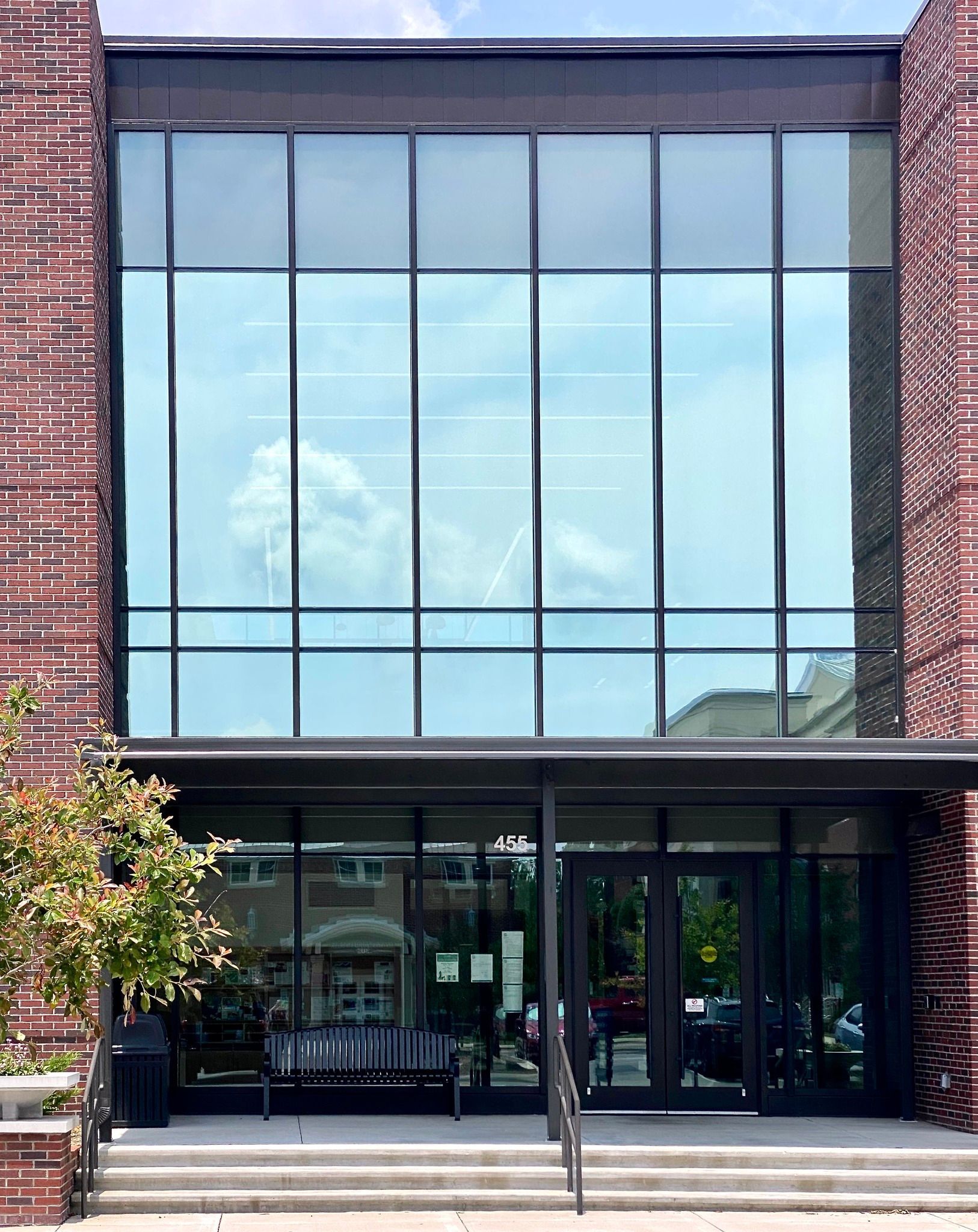 Modern building entrance with large glass windows, brick sides, steps, and a bench outside.