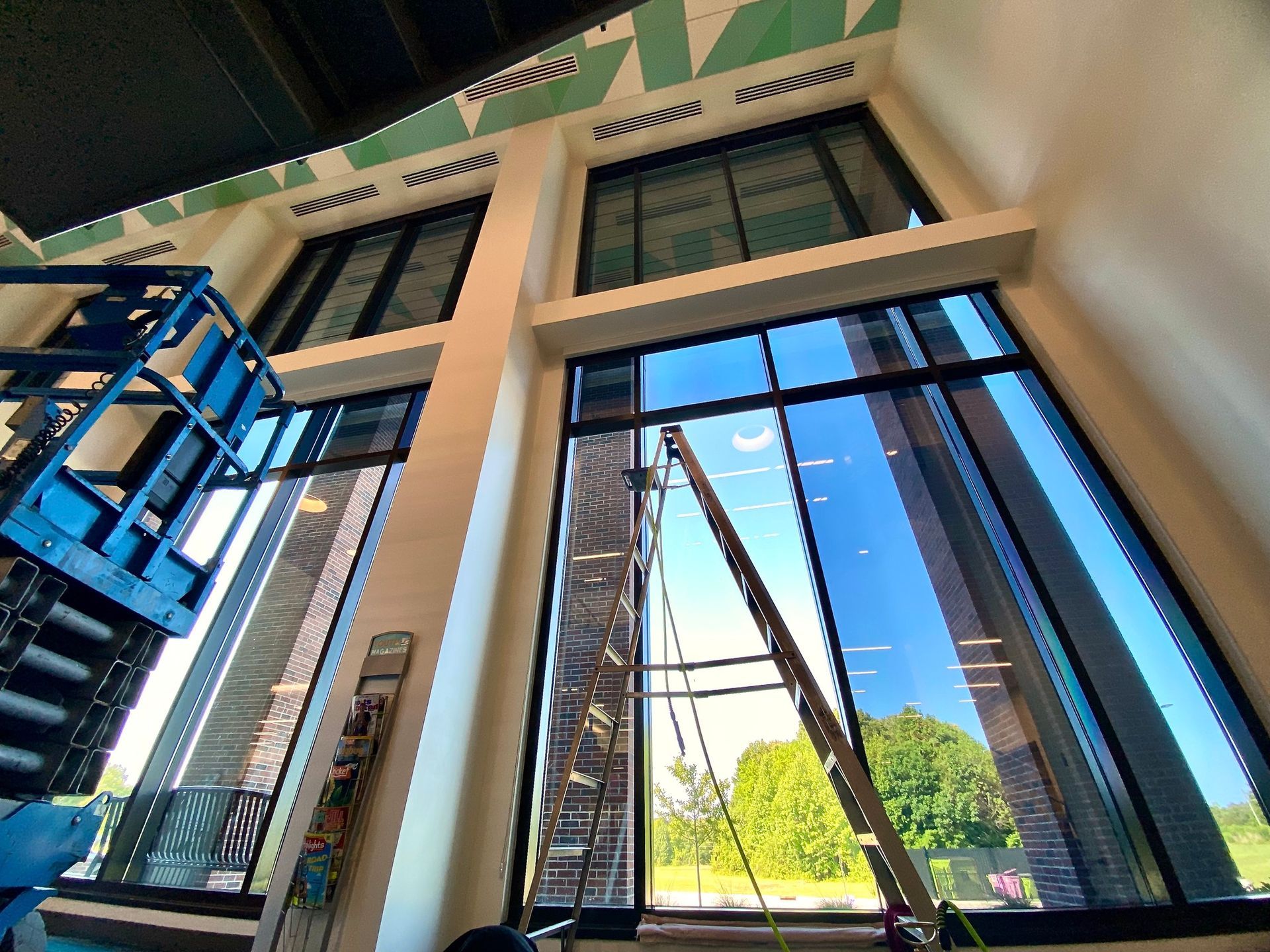 Interior view of tall, floor-to-ceiling windows under construction with a scissor lift and a ladder in the foreground.
