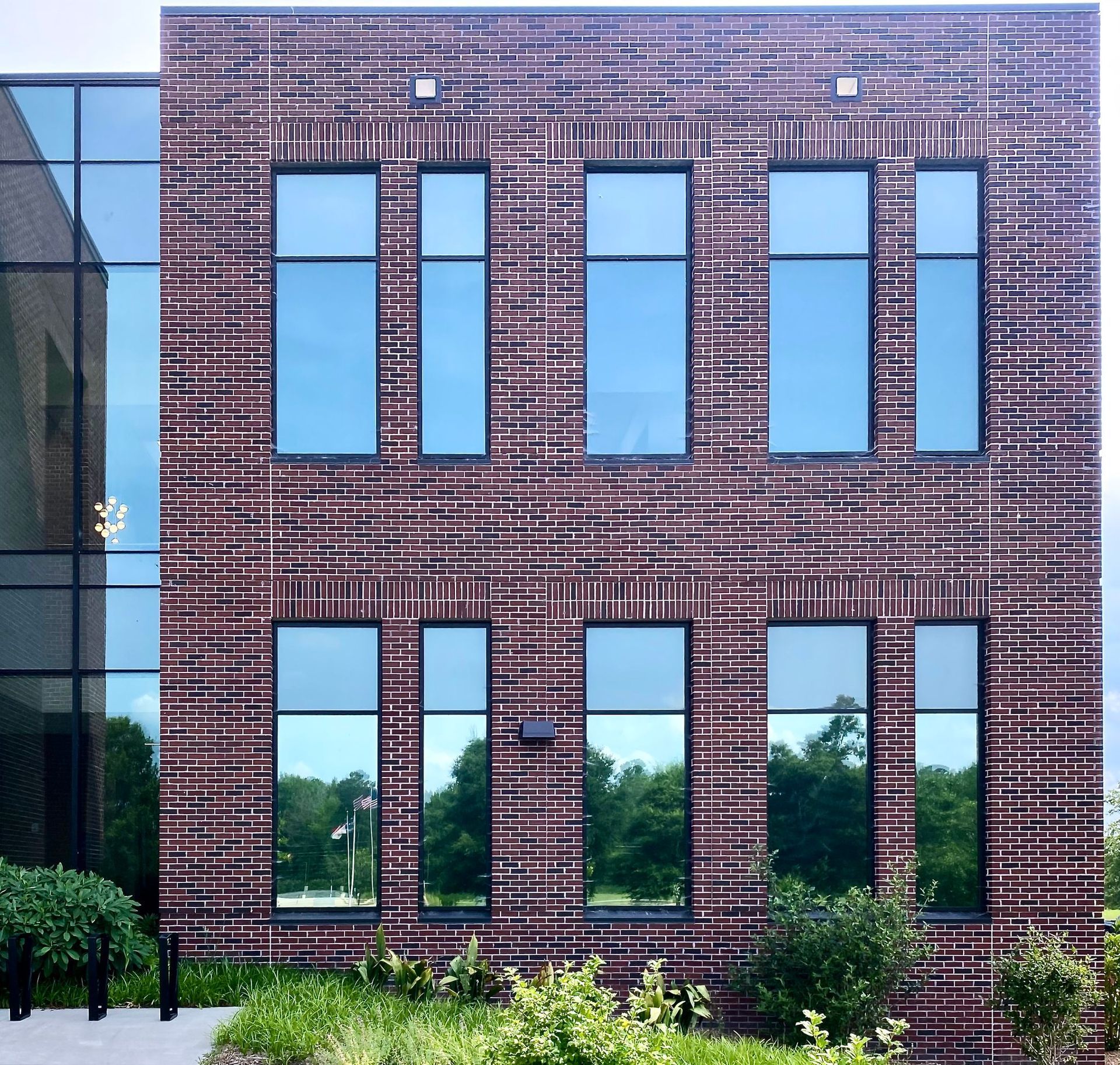 Modern two-story building exterior with dark, textured brick, grid-patterned windows, and green shrubs at the base.