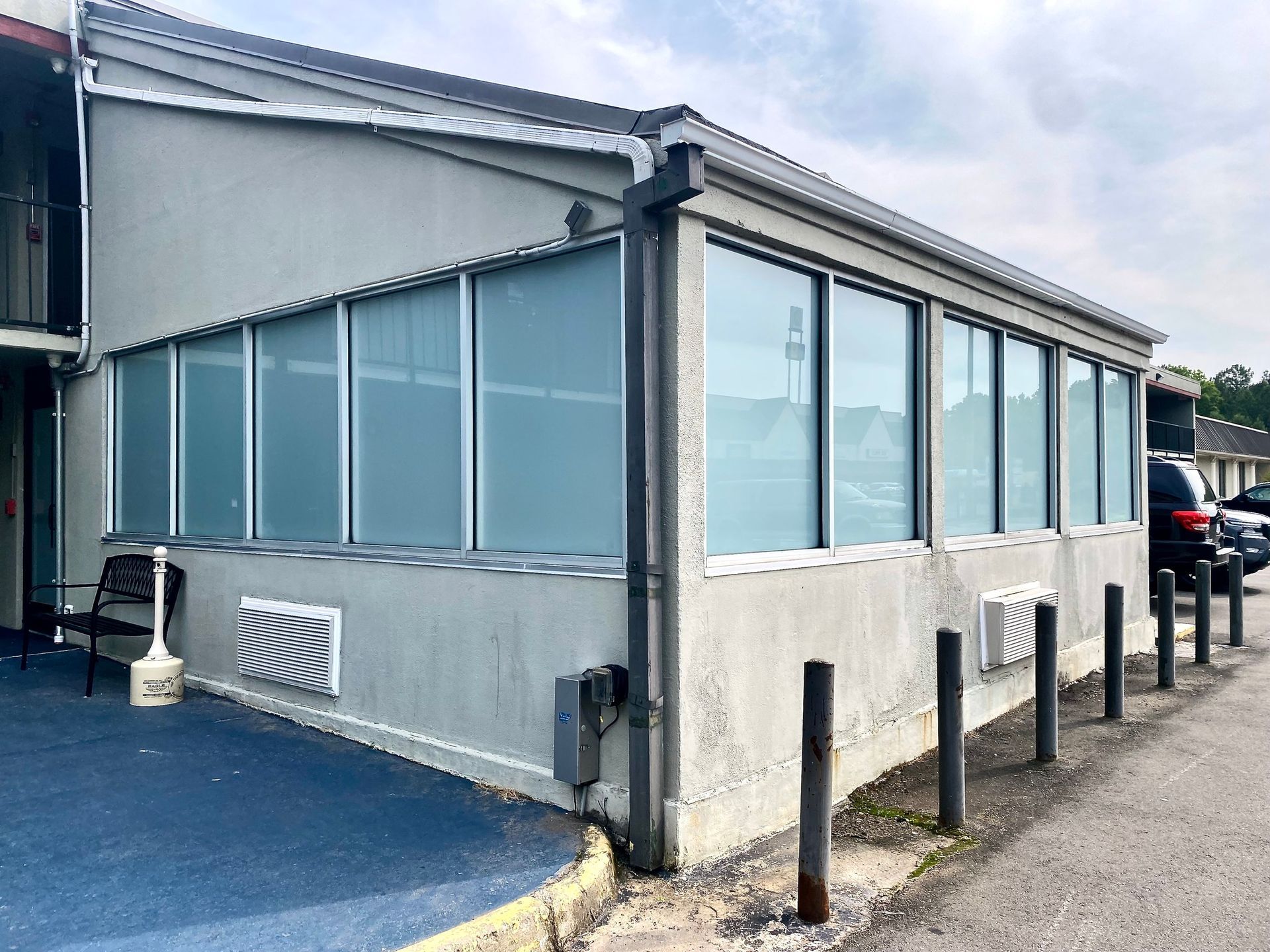 Corner view of a beige building with a row of large, tinted windows, a downspout, and bollards lining the pavement.
