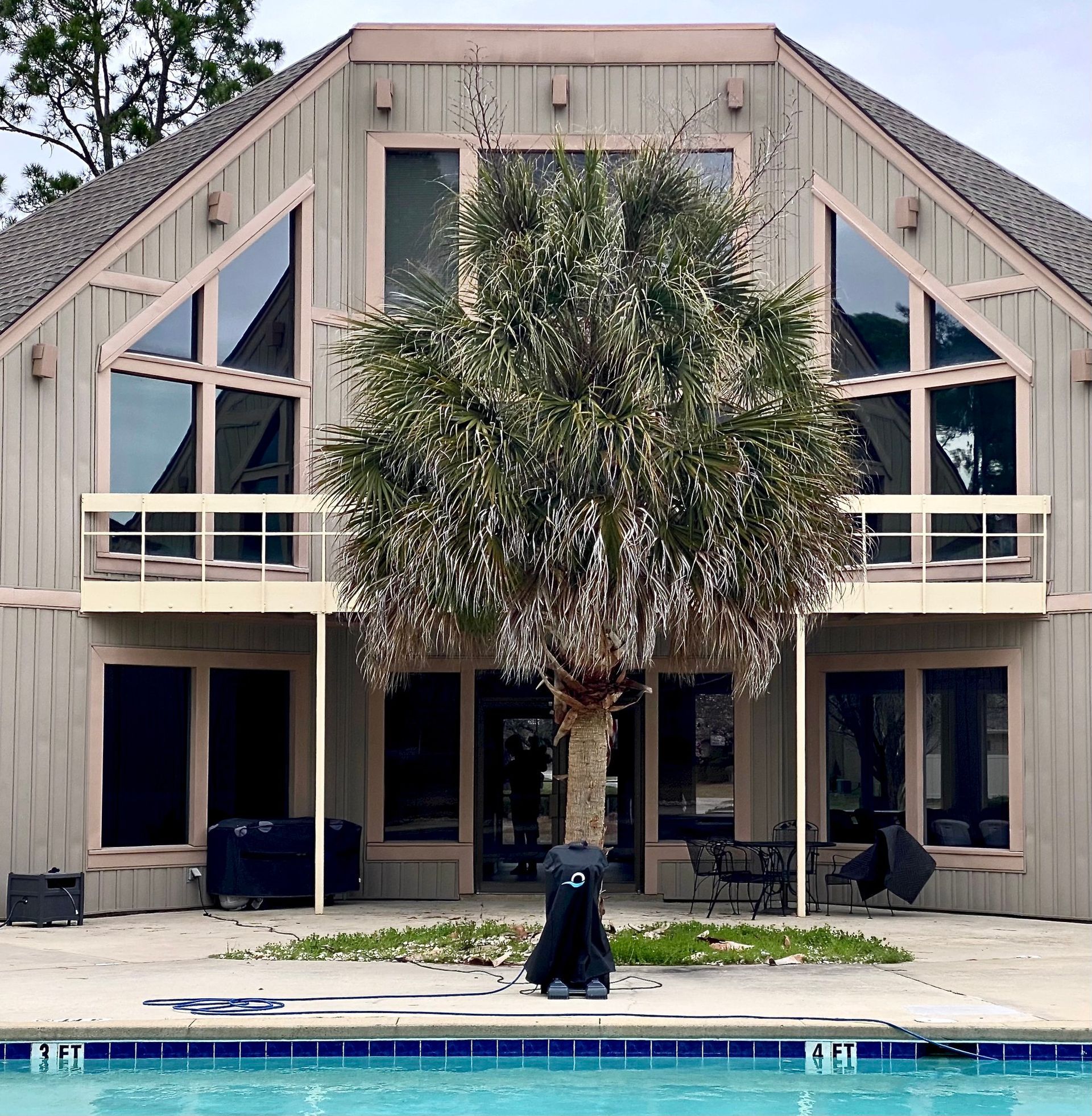A beige, two-story house with a prominent central tree, symmetrical balconies, and a swimming pool in the foreground.