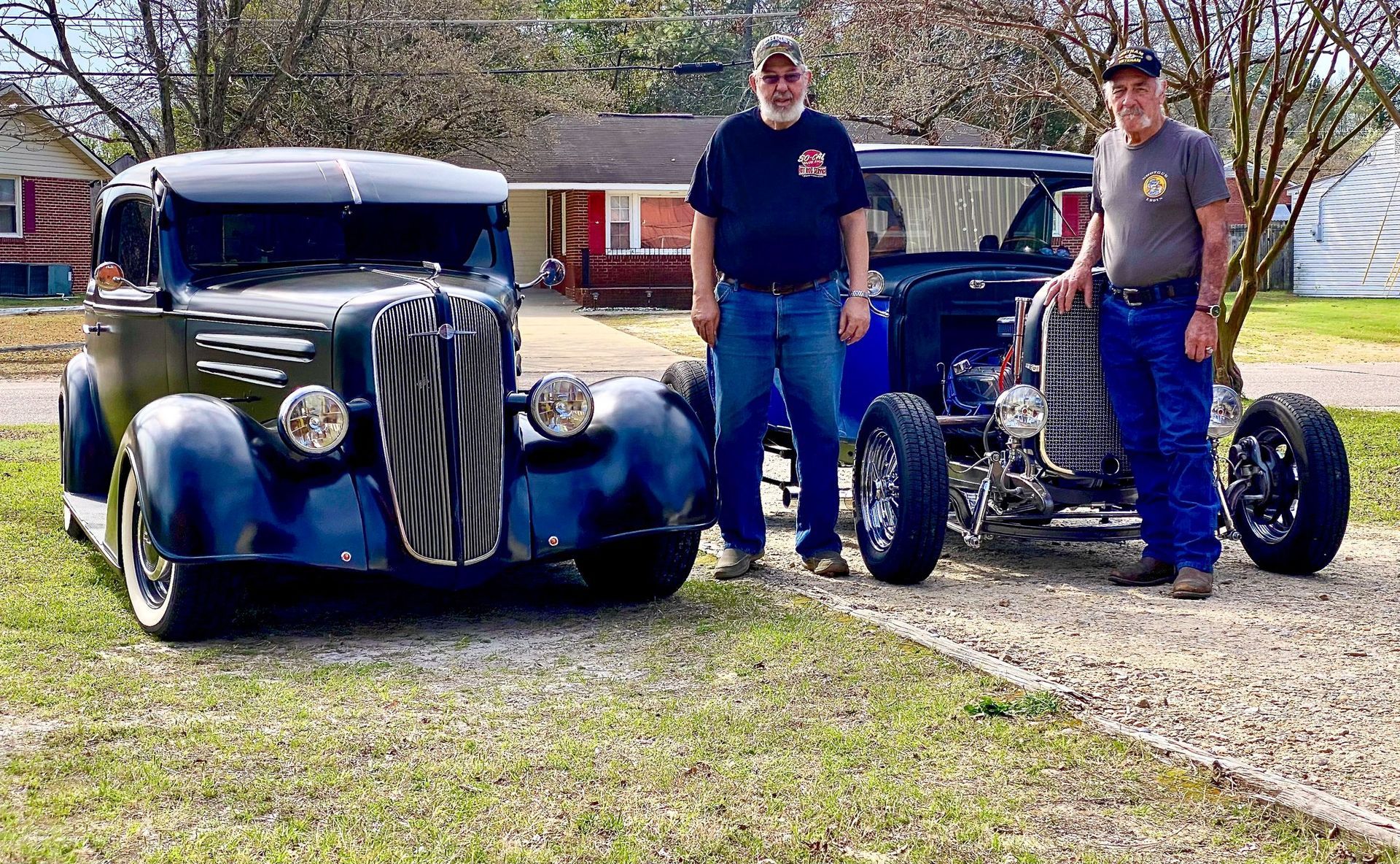 Two people stand in a gravel driveway between a dark vintage car and a blue open-wheel hot rod.