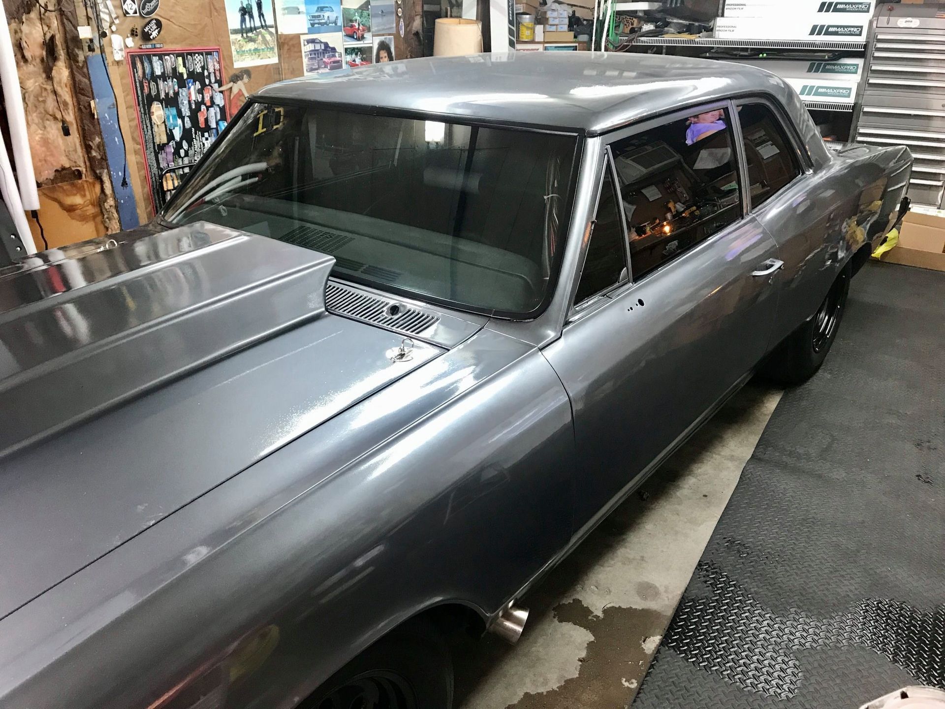 A gray, modified classic muscle car with a large hood scoop, parked inside a cluttered garage workshop.