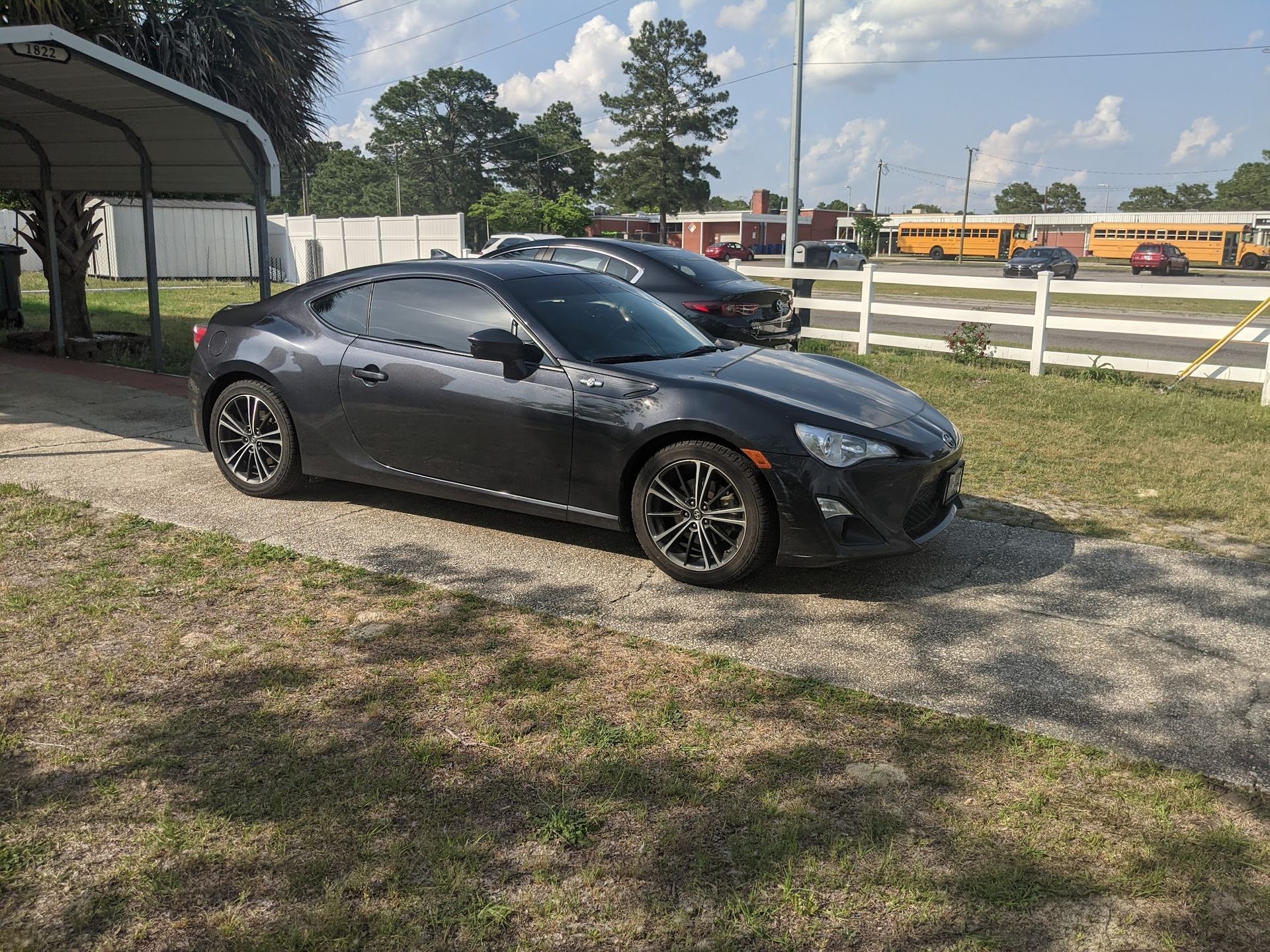 A dark gray Scion FR-S coupe parked on a gravel driveway near a metal carport and a school bus in the background.