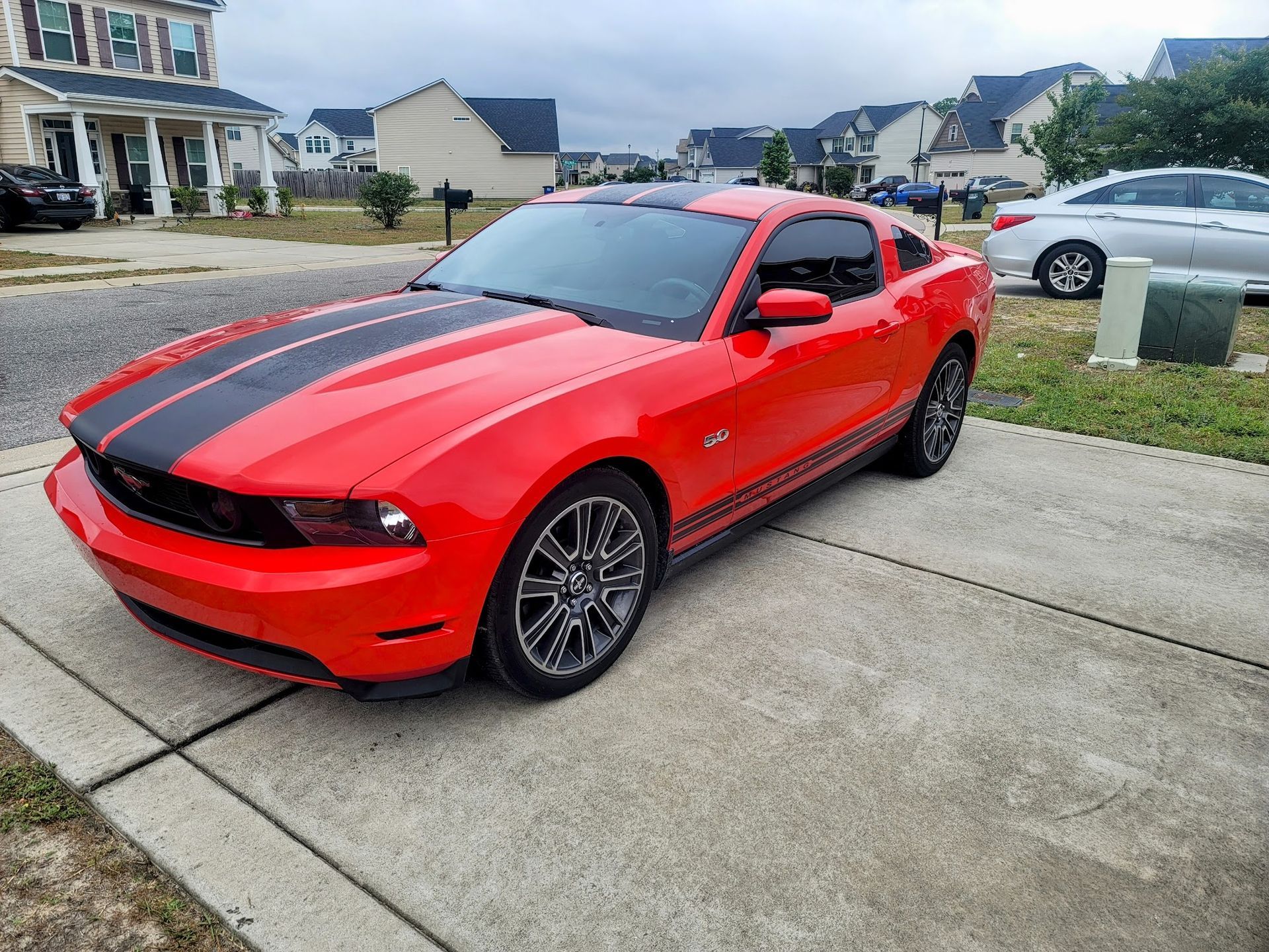 A red Ford Mustang with black racing stripes parked on a concrete driveway in a suburban neighborhood.