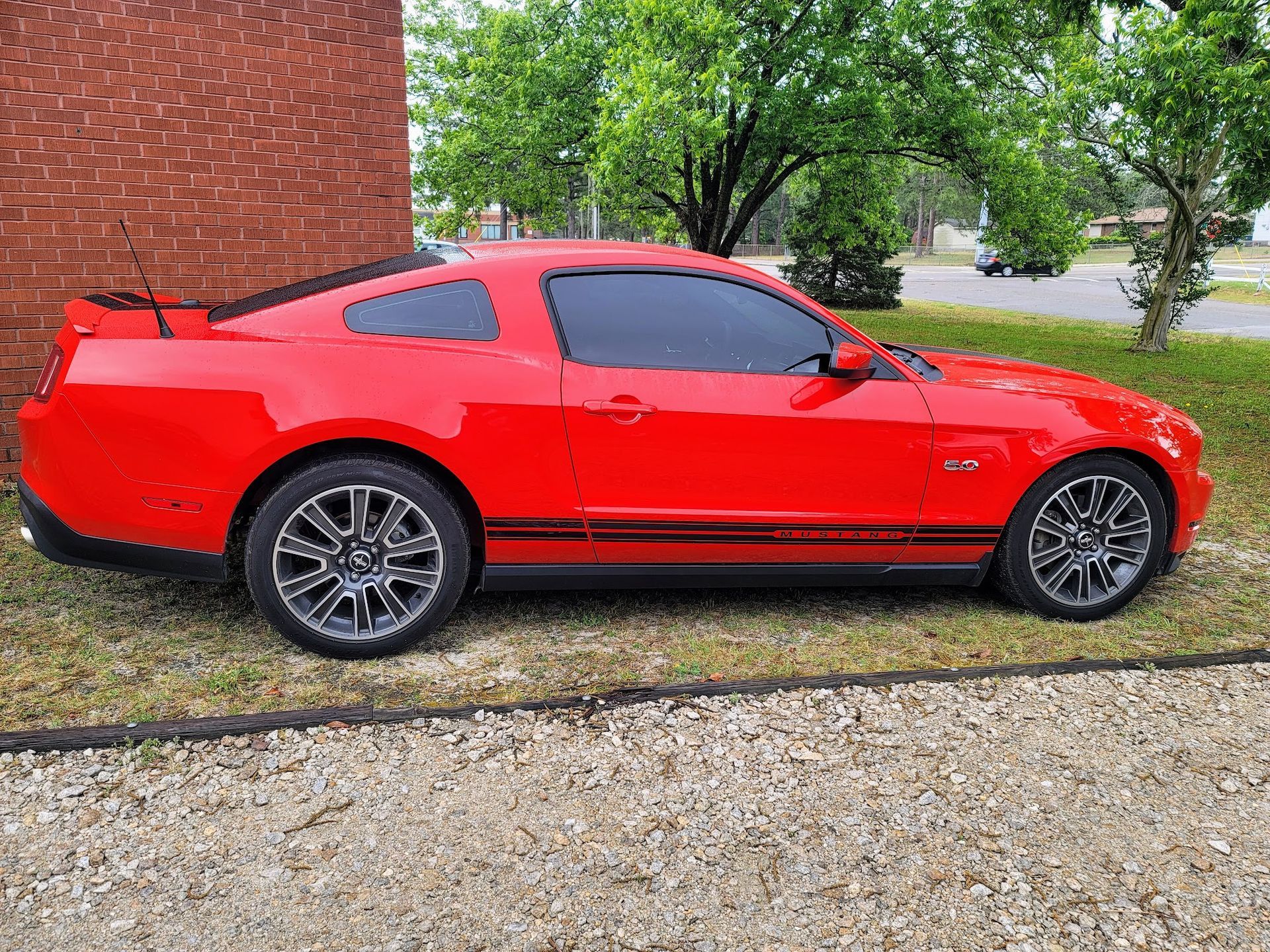 A bright red Ford Mustang coupe parked on a gravel and grass lot next to a brick building.