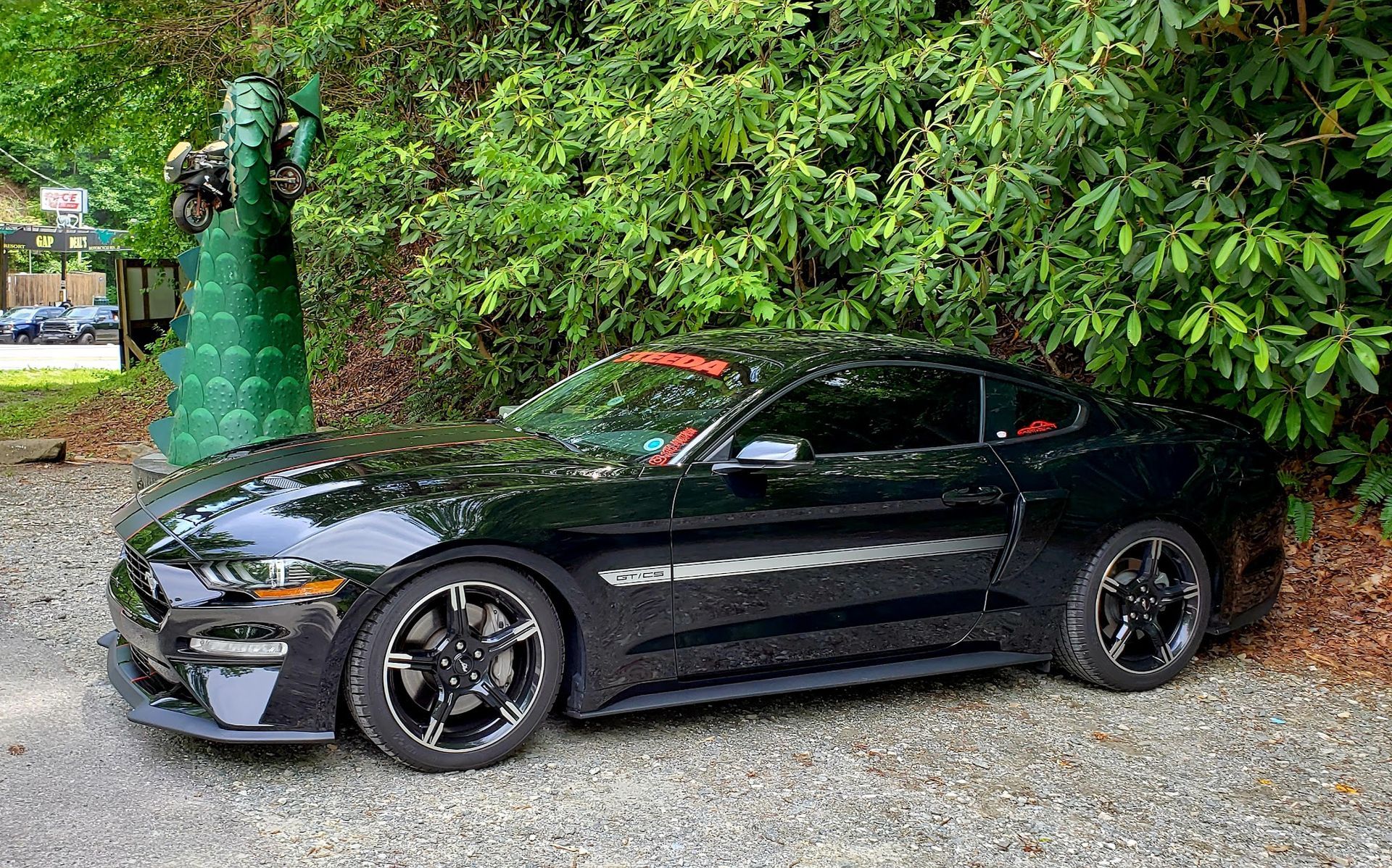A black Ford Mustang parked on a gravel surface next to a decorative green garden statue and leafy trees.