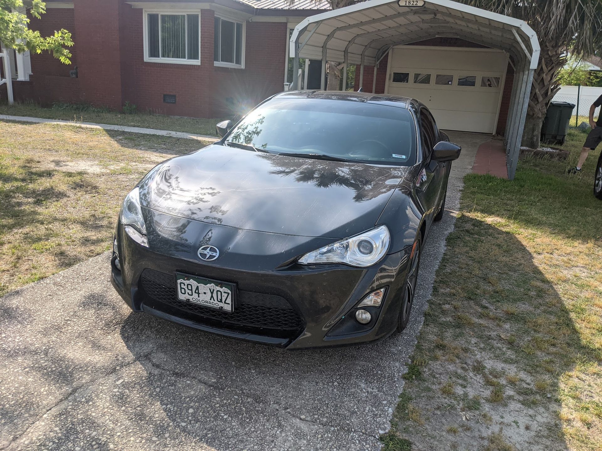 A dark grey Scion FR-S sports car parked on a gravel driveway in front of a house with a metal carport.