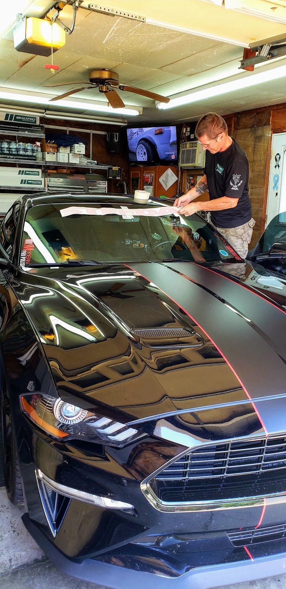A person applying a decal to the top of a black sports car's windshield inside a cluttered garage.