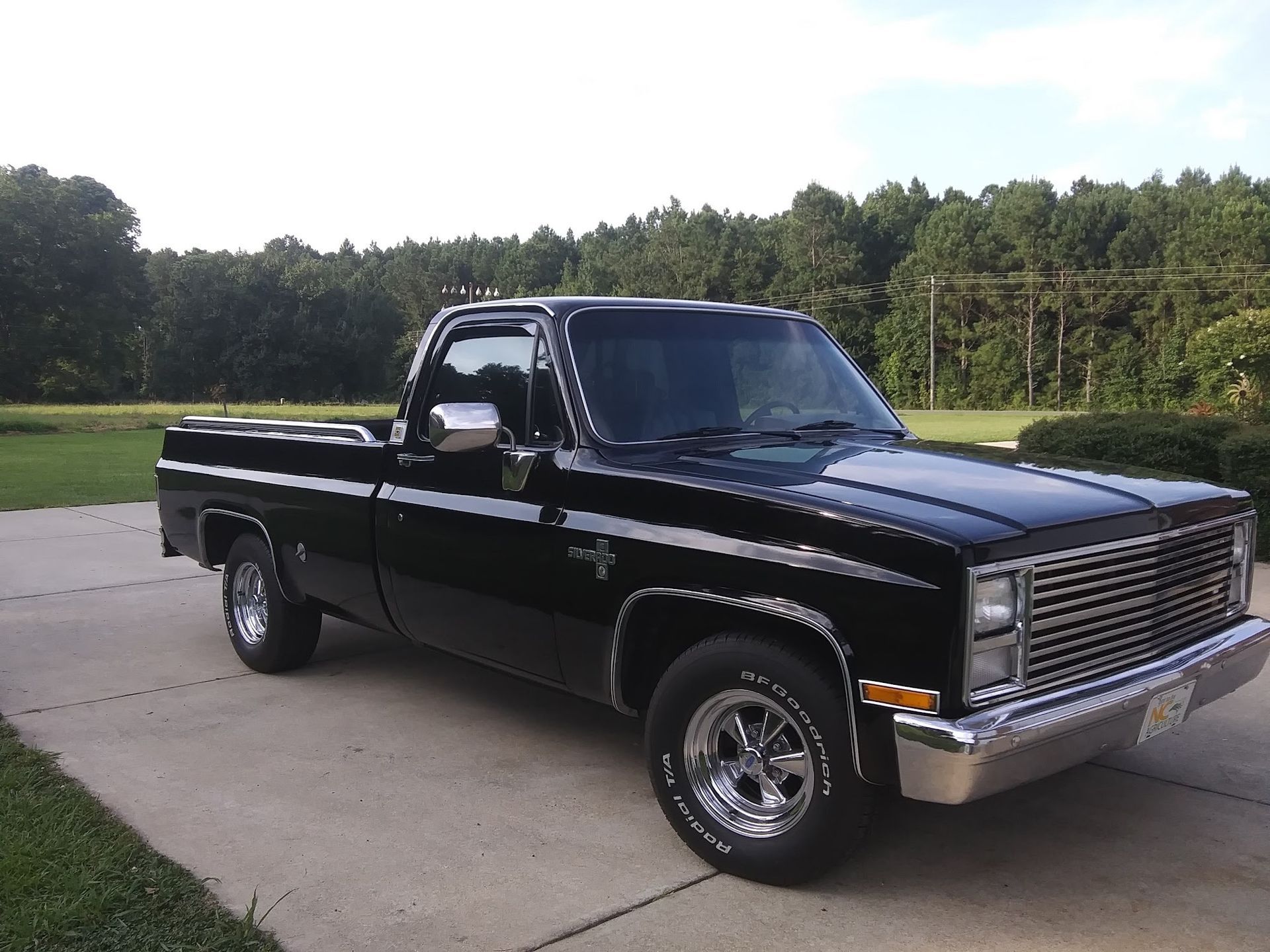 A black vintage Chevrolet C/K pickup truck parked on a concrete driveway with a grassy and wooded background.