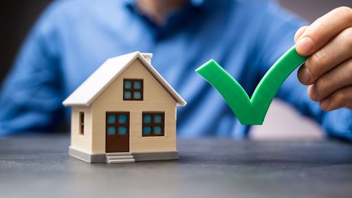 A person wearing a blue shirt is holding a large green checkmark next to a small model of a house placed on a table.