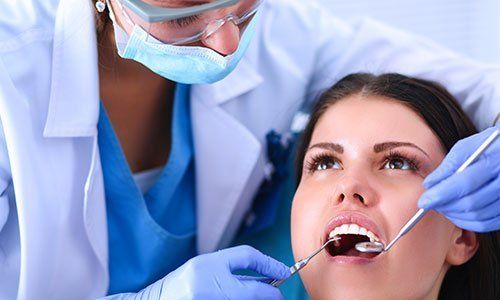 una donna con la bocca aperta sdraiata su un lettino e una dentista che le controlla la bocca con uno specchietto e un rampino