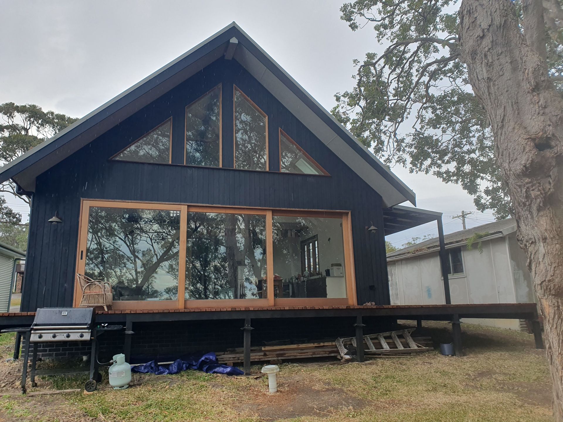 Black cabin with large windows, deck, and a grill on a cloudy day.