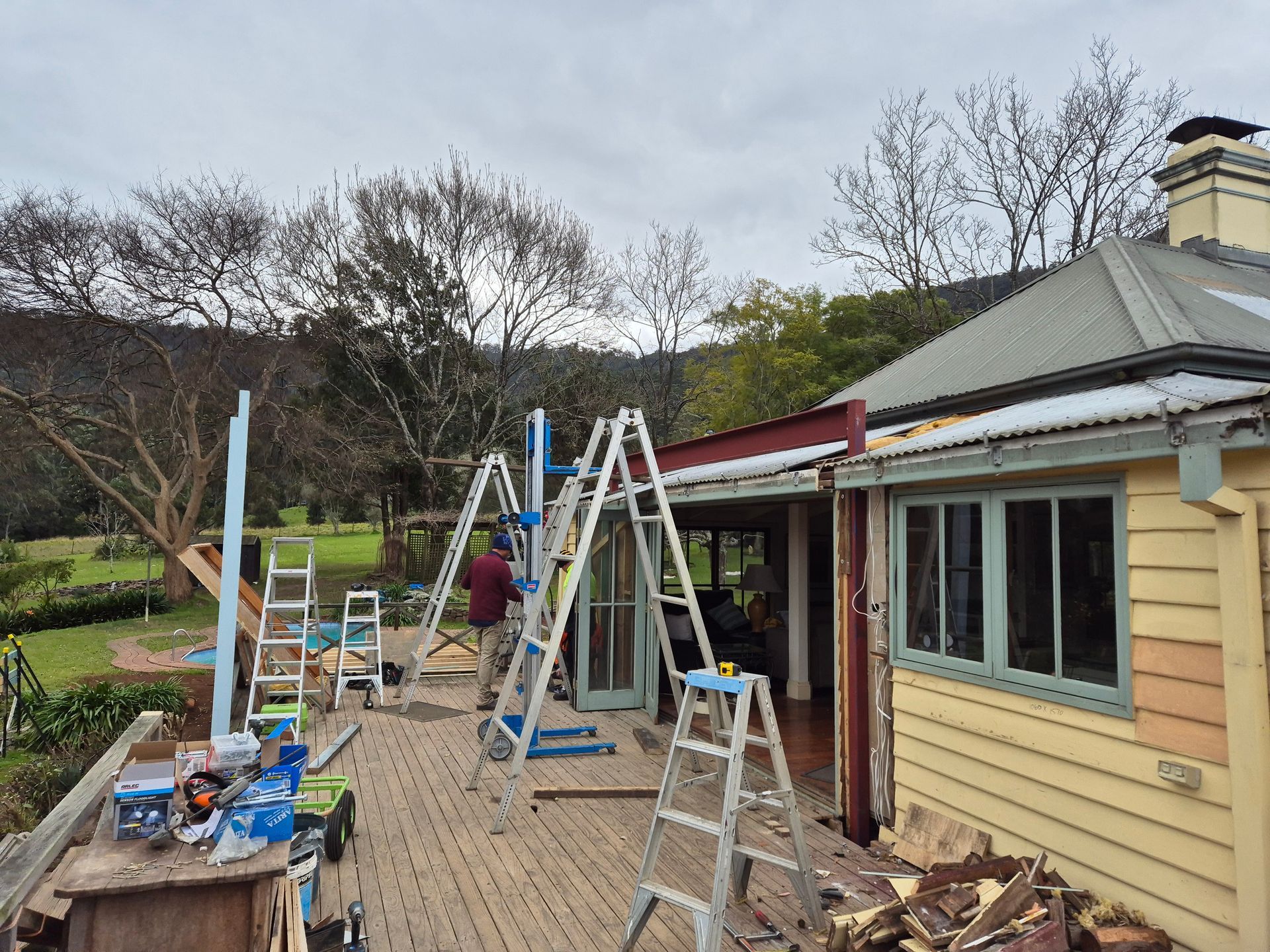 Construction site with workers on ladders rebuilding a patio and house with a rural backdrop.