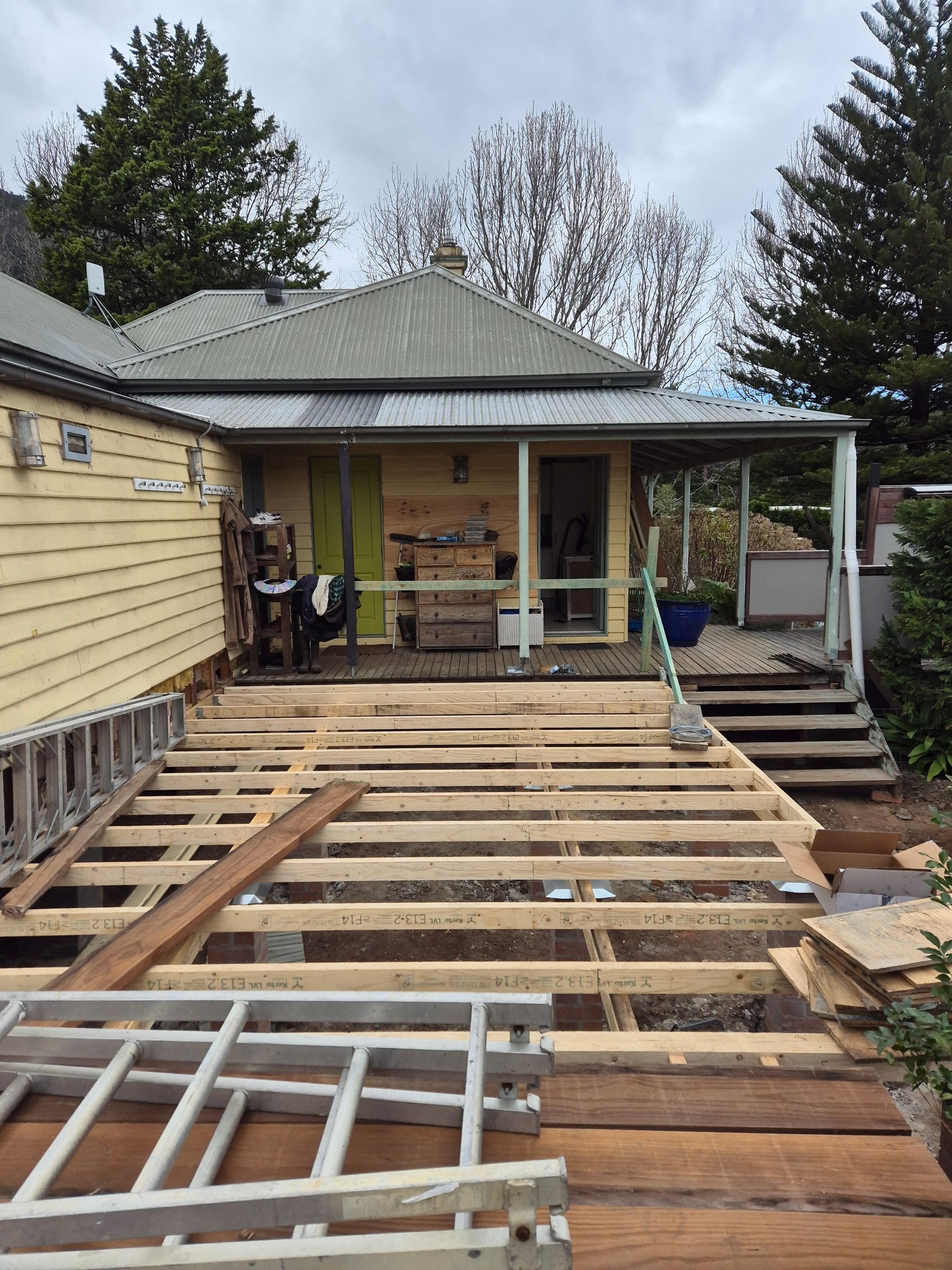 House under construction, wooden deck frame visible, with a yellow house in the background.