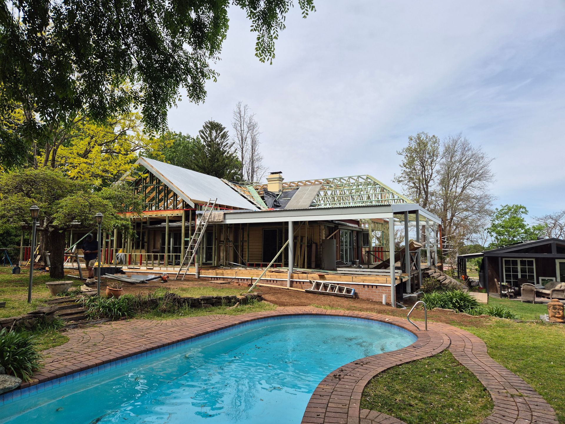 Poolside scene at sunset. Pool surrounded by light stone, with furniture and a house in the background.
