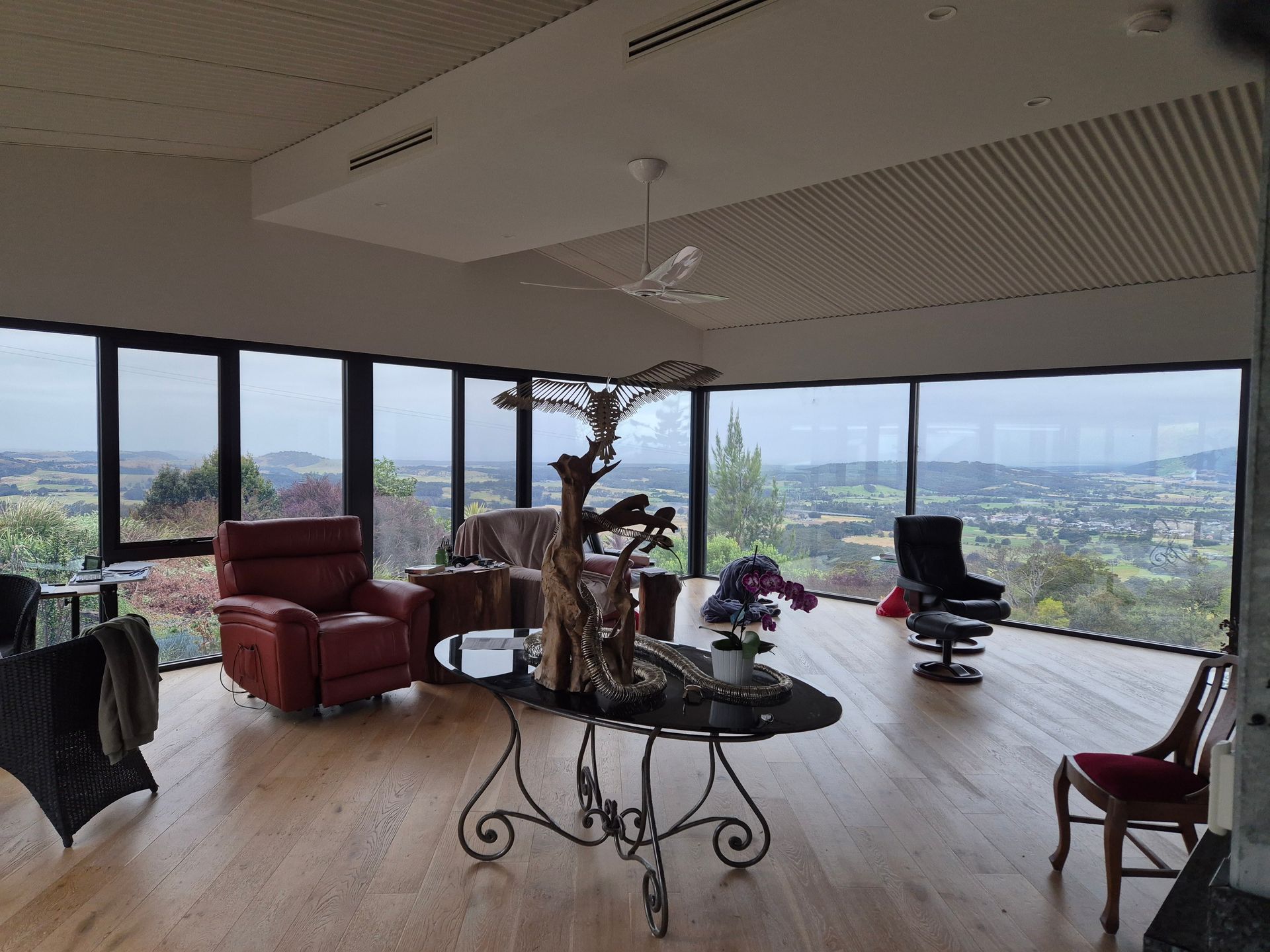 A living room with large windows overlooking a landscape. A decorative fountain, chairs, and a red recliner are present.