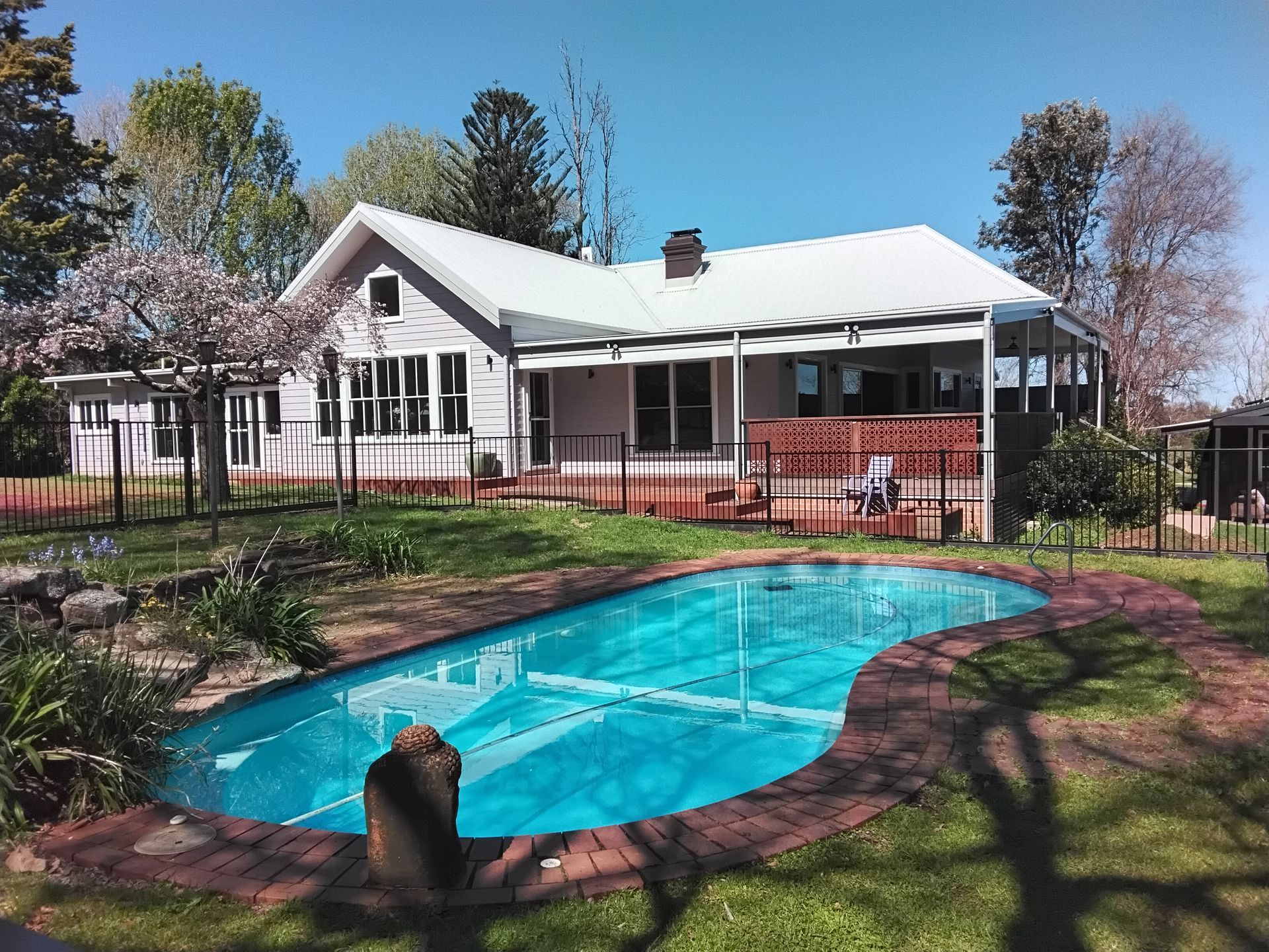 A house with a pool covered by blue tarp, surrounded by a brick walkway and fence, under a sunny sky.