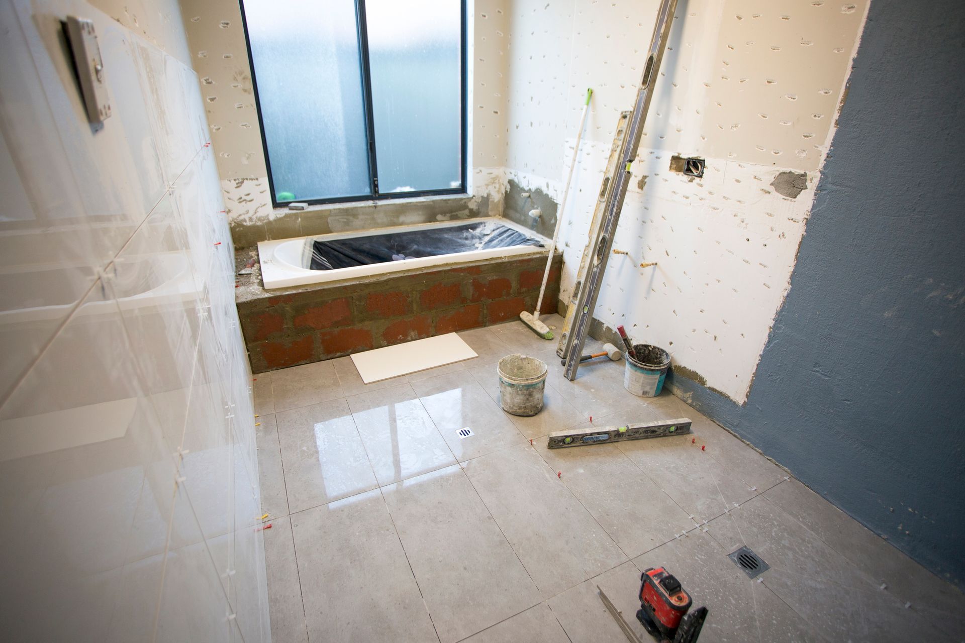 Bathroom under construction, tiled floor, brick tub surround, window, tools scattered.