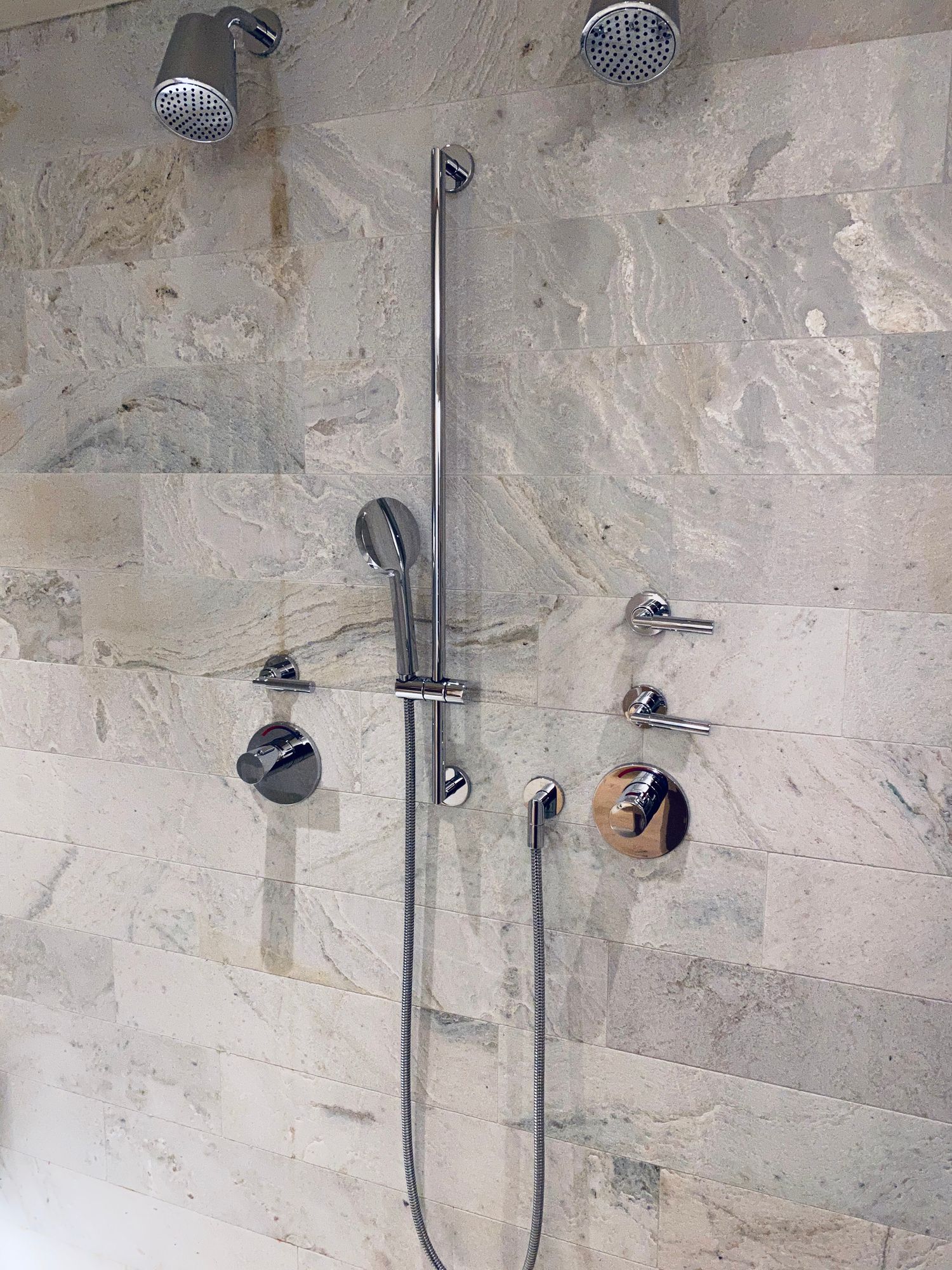 Shower with multiple chrome showerheads and controls on a light stone tile wall.
