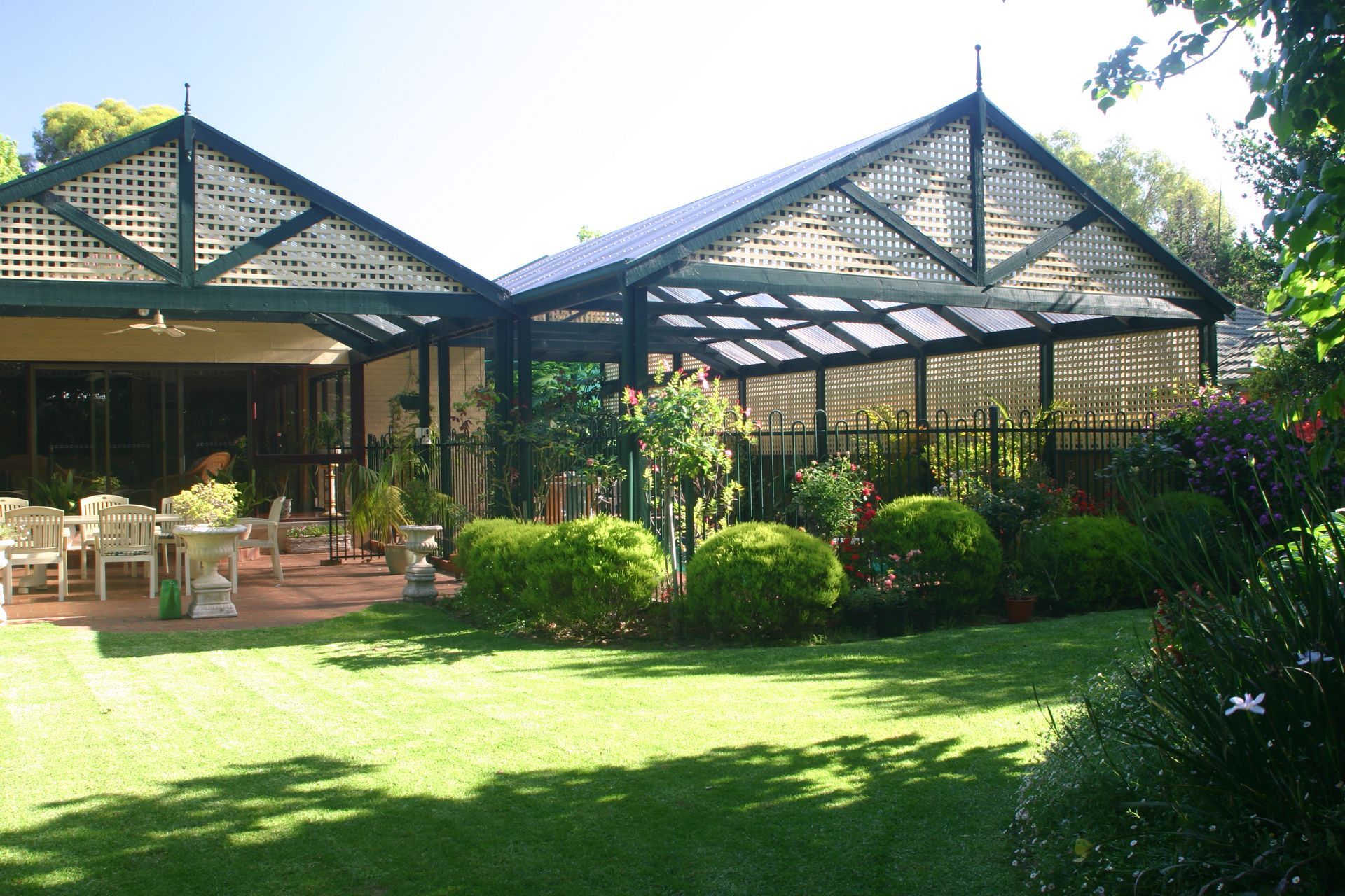 House with a garden, including a lawn, hedges, and outdoor seating under a lattice roof.
