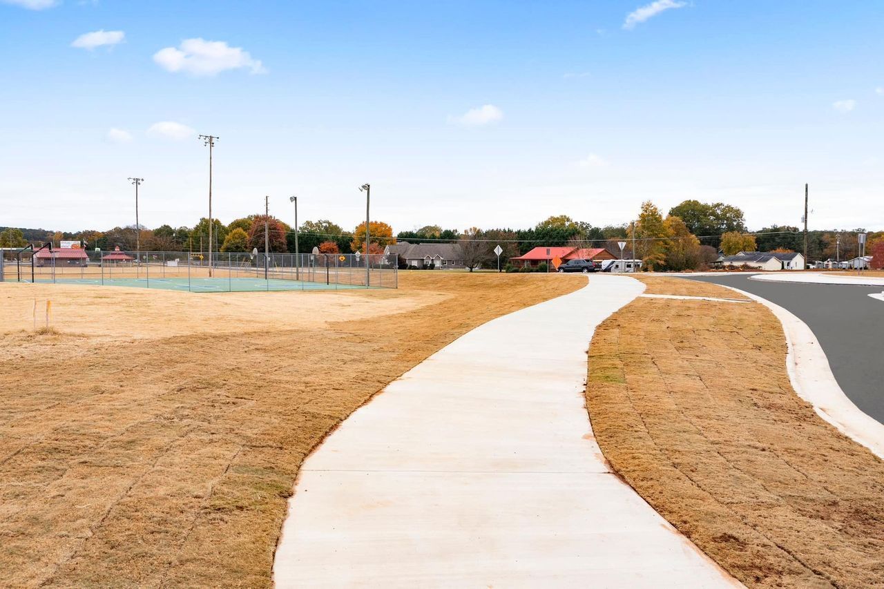 A concrete walkway leading to a field with a tennis court in the background.